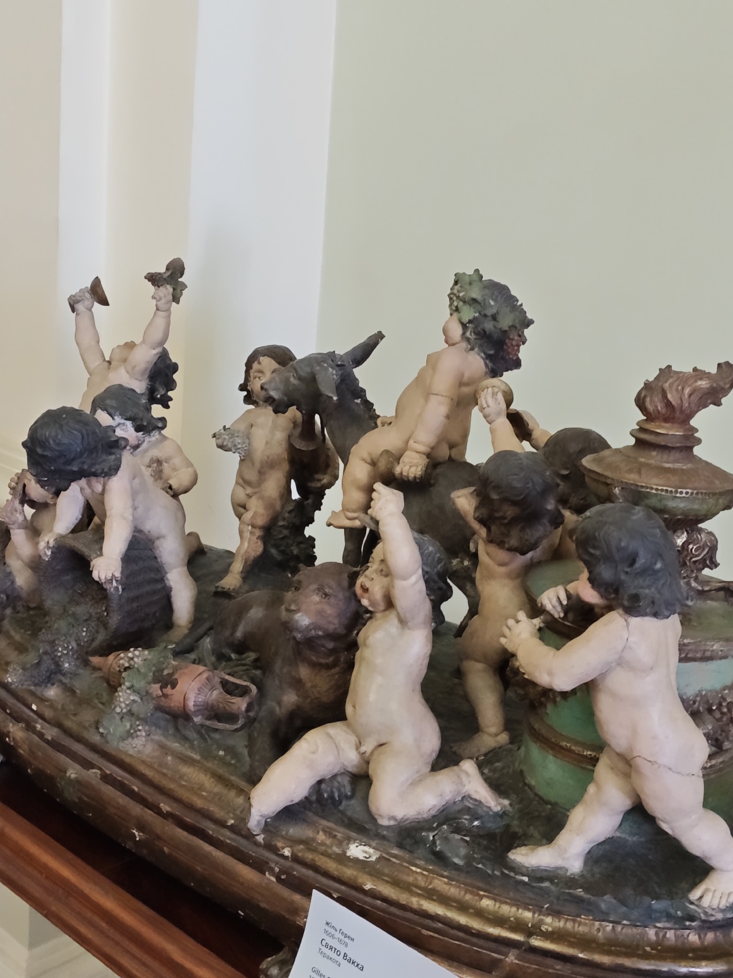 This image depicts a detailed terracotta sculpture featuring a group of cherubic figures, likely part of a larger decorative piece or fountain. The figures are in various dynamic poses, some climbing, some reaching, and one wearing a wreath of grapes. There are also animalistic elements, including what appears to be a dog or lion-like creature and the head of a donkey or horse. The sculpture is set on a base that resembles a rocky or earthy terrain, with some green foliage and ceramic fragments. The sculpture is displayed indoors, with a plain, light-colored wall as the background. A label with Cyrillic text is visible at the bottom right corner, suggesting the location might be in Lviv, Ukraine, possibly at the Lviv National Art Gallery, where similar Baroque sculptures are housed.