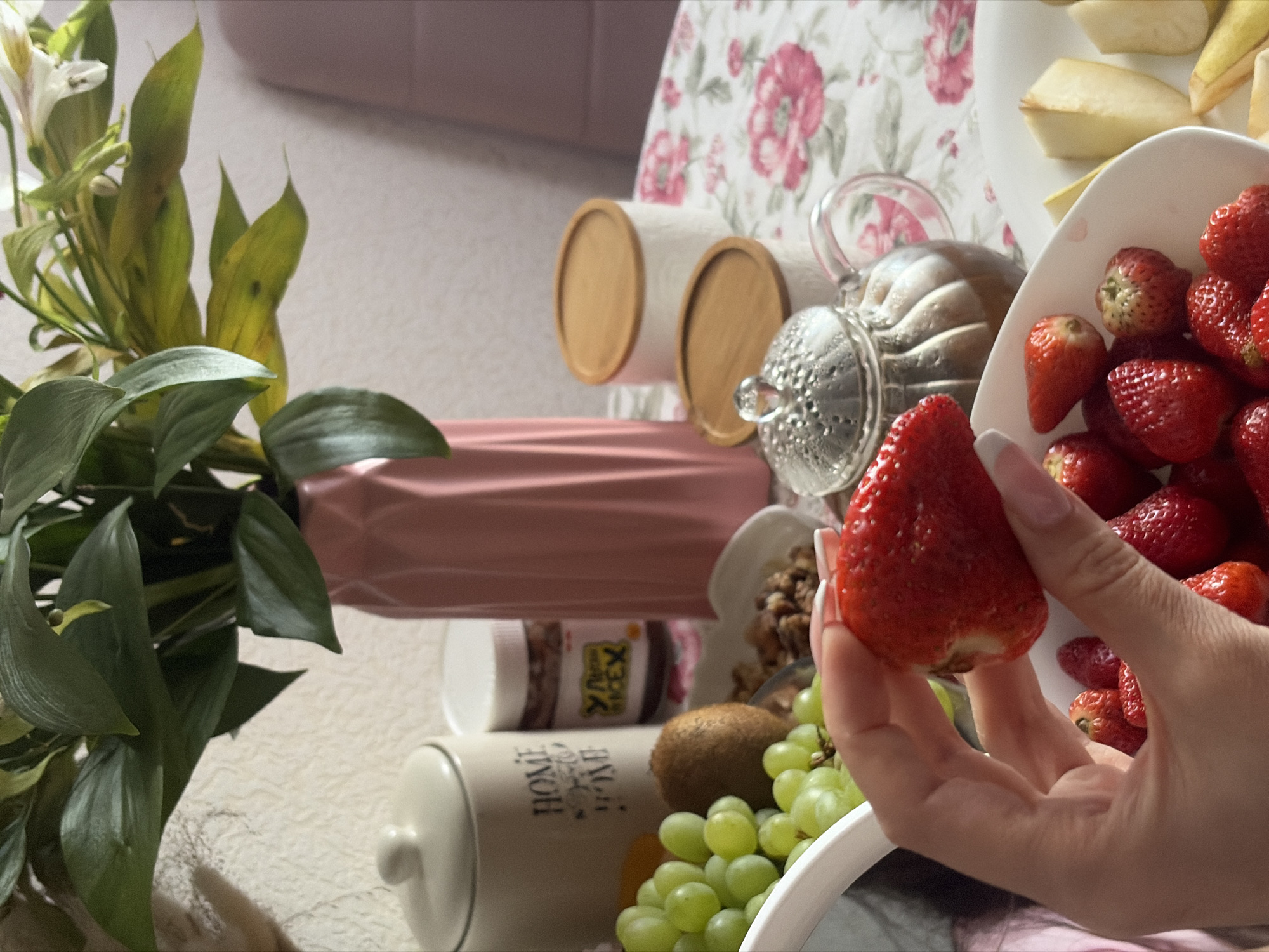 The image shows a still life arrangement with a focus on food and domestic objects. A hand holds a large strawberry in the foreground, with a bowl of strawberries and other fruits, including grapes, visible within the bowl. To the left of the bowl is a kiwi and a jar or canister with the word "HOME" visible on it. Behind these items is a jar with the word "Spicemex" visible on it. Above the jars is a horizontal pink vase or decorative object.
Behind this arrangement, there are two white canisters with wooden lids. To the right of these, there is a glass teapot with a metal strainer. The background consists of a fabric with a floral pattern. To the left is a bouquet of flowers with green leaves.
There is no indication of the location in the city of Saint Petersburg, Russia, within the image.