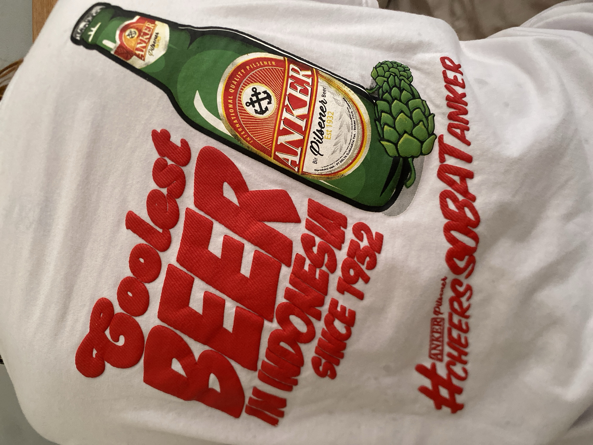 The image shows a white t-shirt with a large graphic of a green "ANKER Pilsener" beer bottle. Next to the bottle, there's a green hop. Below the bottle is the text "Boolest BEER IN INDONESIA SINCE 1932" and "#CHEERS SOBATANKER" in red. The text indicates the beer is available in Indonesia and the location is the city of North Kuta in Indonesia.