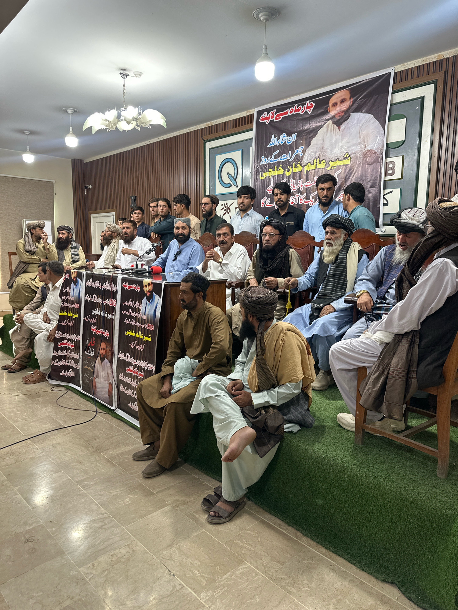 A group of men, some wearing traditional Pashtun attire and turbans, are seated and standing in what appears to be an indoor event. Behind them, a large banner with images and text in Arabic script is displayed on a wooden paneled wall. Some of the men are seated at a long table covered with green carpet, facing forward, and a few are speaking into microphones. The overall setting suggests a press conference or a political gathering. The presence of a banner with a portrait of a man implies the event is related to this individual. The floor is tiled, and a chandelier hangs from the ceiling.