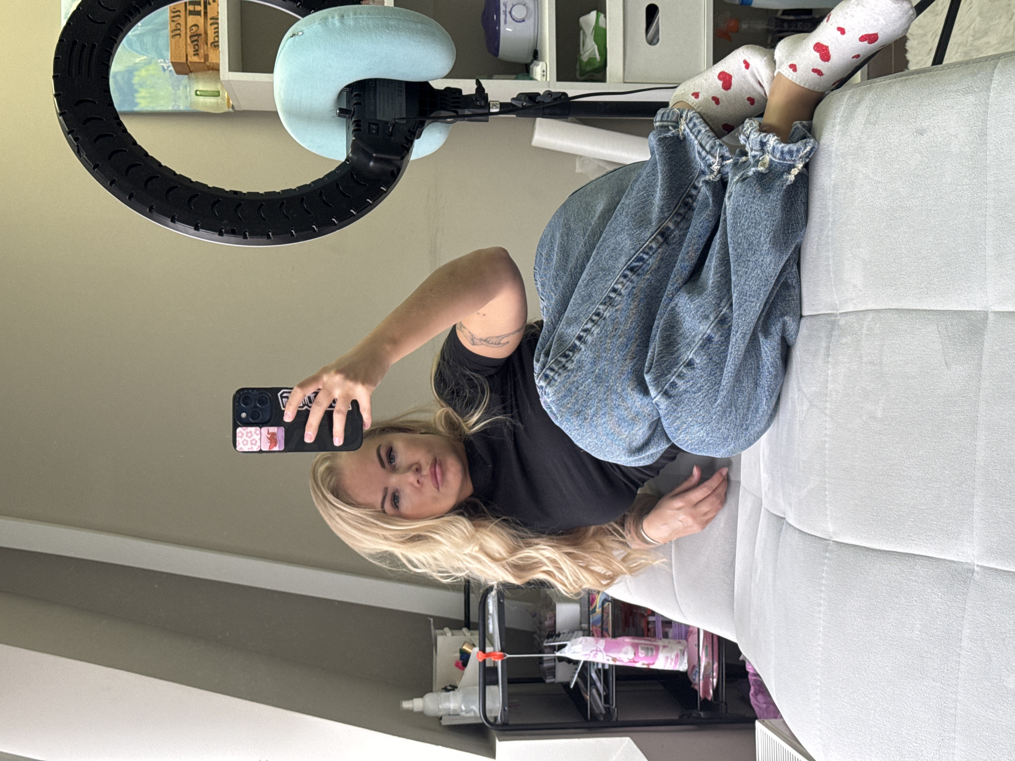 A woman is taking a selfie while lying on a light gray couch. She has long blonde hair and is wearing a black shirt and blue jeans. Her feet are clad in white socks with red hearts. Behind her is a ring light attached to a stand with a blue headrest. Various items are arranged on a white shelf in the background. The scene is indoors.