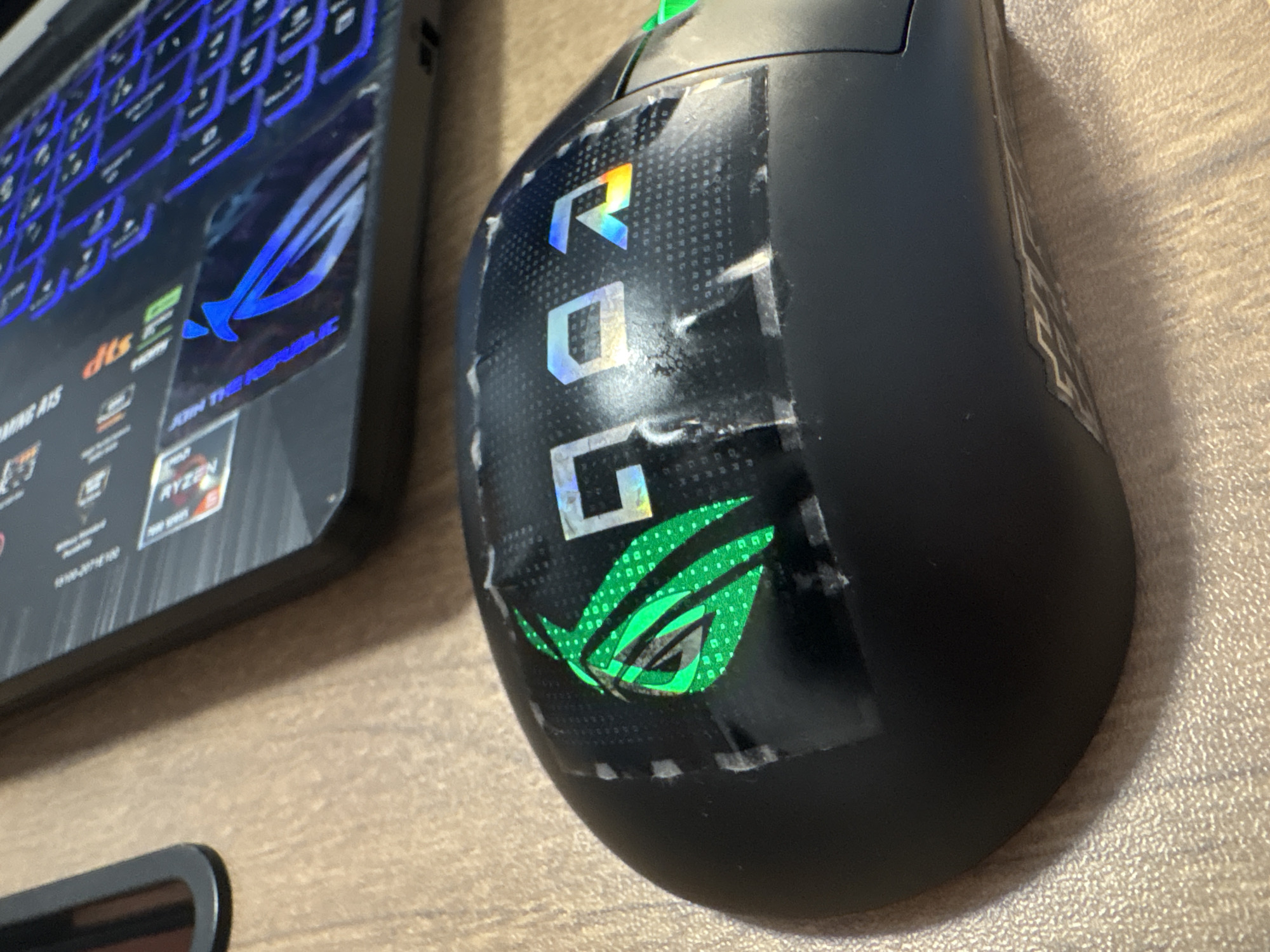 This image shows a close-up of a black gaming mouse adorned with stickers. The stickers feature the "ROG" logo and the Republic of Gamers eye emblem, both with a holographic, iridescent sheen that shifts in color with the light. The "ROG" letters are placed vertically on the mouse, with the "R" at the top, followed by "O" and "G" below. The ROG eye emblem is prominently displayed below the letters. Some parts of the stickers appear to be peeling or lifting, suggesting they have been applied for some time. The mouse is positioned on a light brown, textured surface, possibly a desk or table. To the left of the mouse, a portion of a laptop is visible. The laptop has a black casing and a keyboard with blue, backlit keys. A prominent blue ROG logo is also visible on the laptop's palm rest. The overall scene suggests a gaming setup or a workspace related to gaming. There are no people visible in the image. The lighting appears to be from a natural source, possibly daylight, casting soft shadows. No specific time of day or weather conditions can be determined. The visible text includes "ROG" on the stickers and on the laptop, as well as what appears to be "JOIN THE REPUBLIC" below the ROG logo on the laptop. There are also other smaller text elements on the laptop, some of which are partially obscured. The context provided states "Małkowo, Poland," which is a geographical location, but there are no specific visual cues in the image that confirm this location.