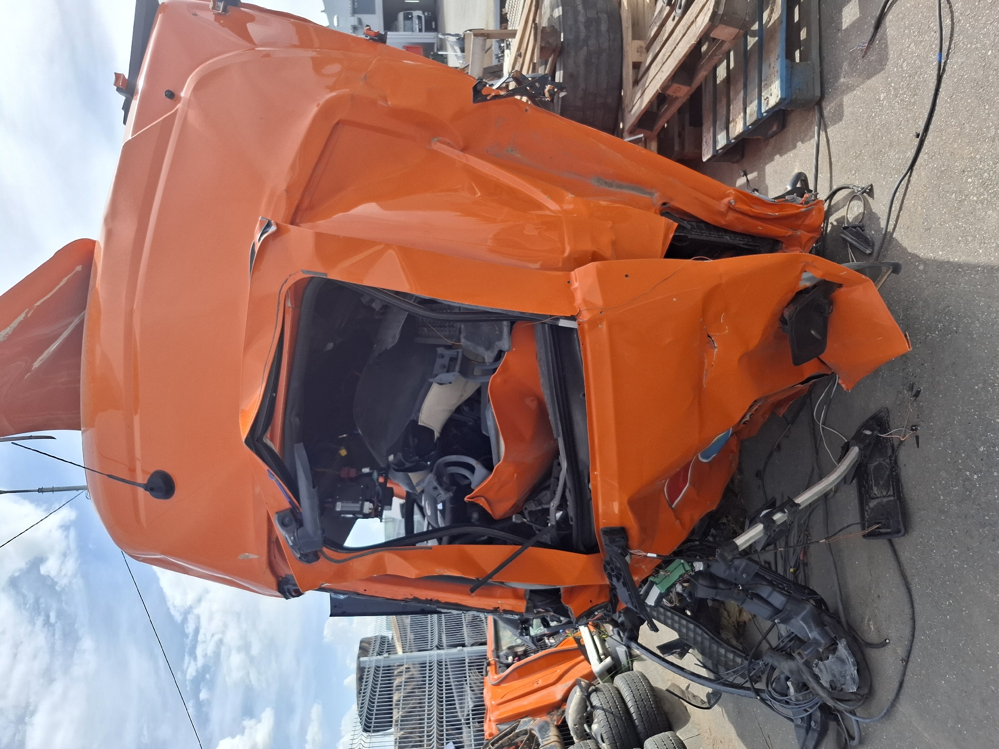 The image depicts a severely damaged orange truck cab. The cab is significantly deformed, with crumpled metal and exposed interior components. The scene appears to be an outdoor storage area, with pallets and other truck parts visible in the background. There is no visible signage indicating the location in Izhevsk, Russia. There are no visible people. The focus is on the damage to the truck cab.