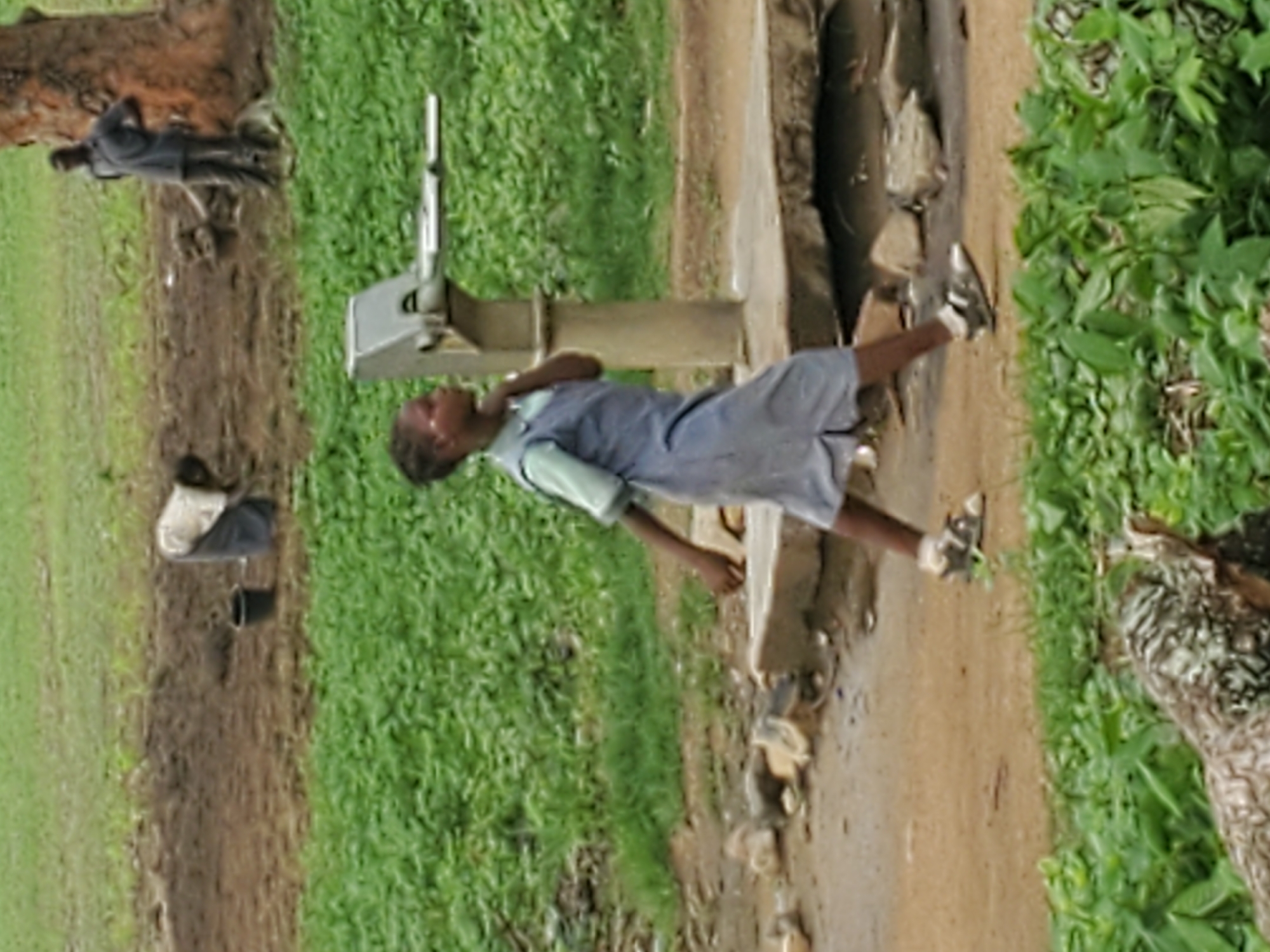 A young girl in a blue dress and white undershirt walks past a water pump in Kabwir, Nigeria. She is walking from right to left, and her left hand is touching her chin. Behind her, a man in a gray shirt stands next to a tree, and further in the background, a person in a white and gray outfit bends over a dirt patch with a black bucket. Green foliage surrounds the scene.