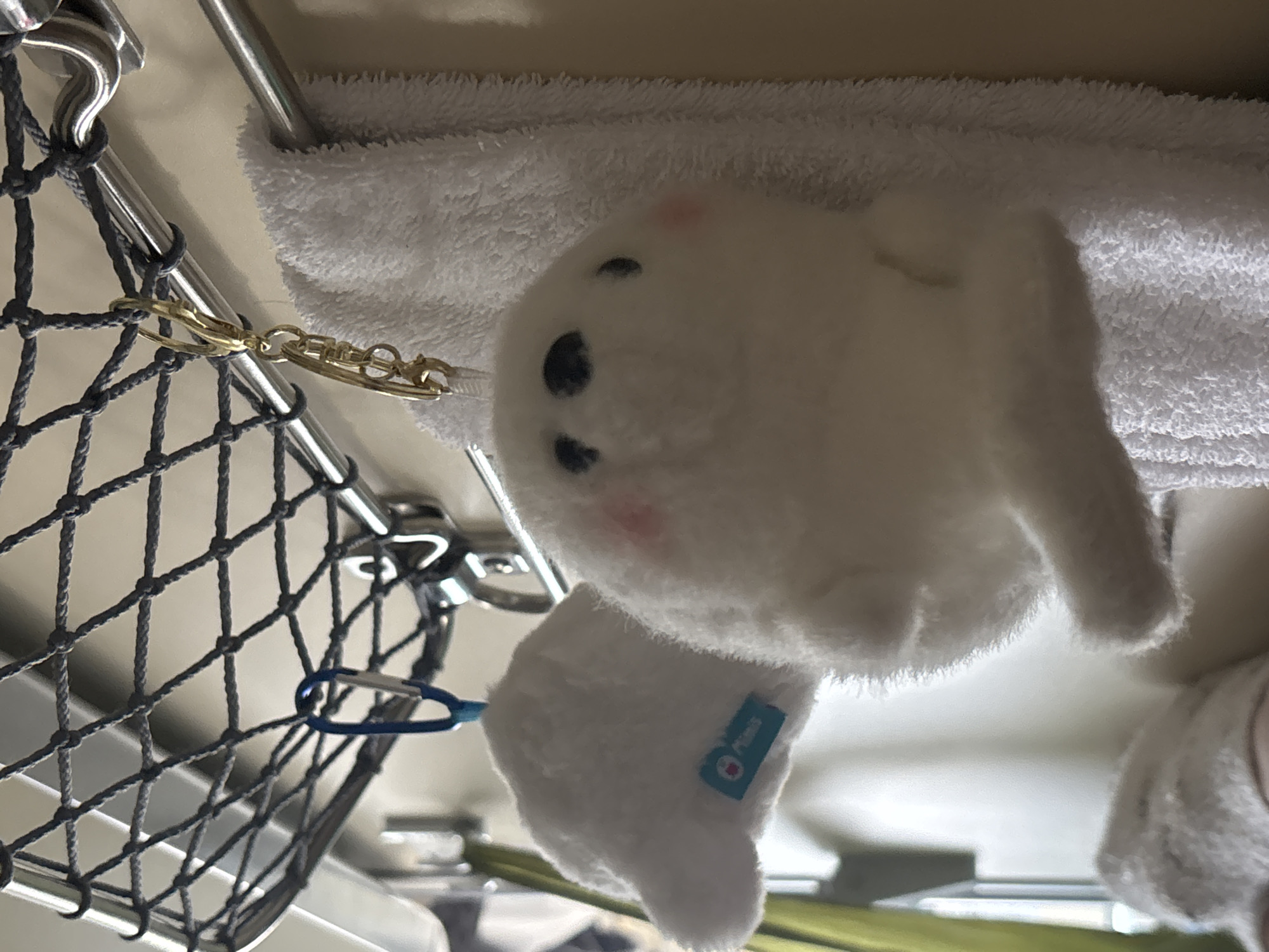 The image depicts an interior scene, characteristic of a train compartment, located in Kalachinsk, Russia. A white, round-bodied plush toy with black eyes and light pink cheek accents is centrally positioned, suspended by a gold-colored keyring from a polished metal rod. This rod forms part of an overhead storage system, which includes a tightly woven grey net directly above. To the left, a second, partially visible white plush toy hangs from a blue carabiner and displays a small light-blue tag with "PRIMUS" in white text. Below the main plush toy, a white terrycloth towel is folded and draped over a lower, parallel metal rail. The background consists of a plain, light-toned wall, with a vertical strip of green fabric or curtain visible on the far left side of the frame.