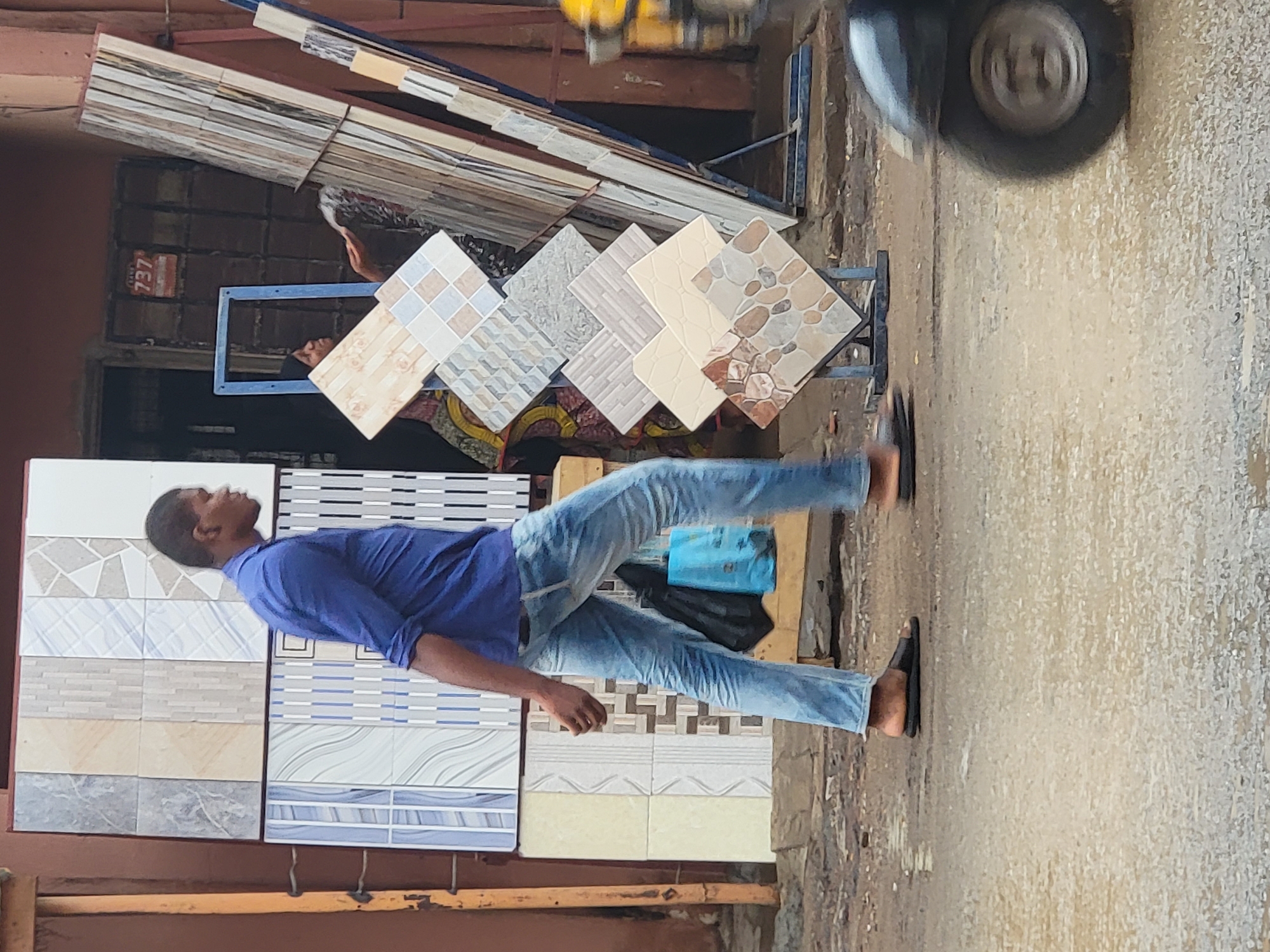 A man in blue shirt and jeans walks past tile samples displayed for sale in Maiburiji, Nigeria. The tiles are arranged in rows and columns, with various patterns and colors. A vehicle tire is visible in the upper right corner of the frame. The ground is wet.