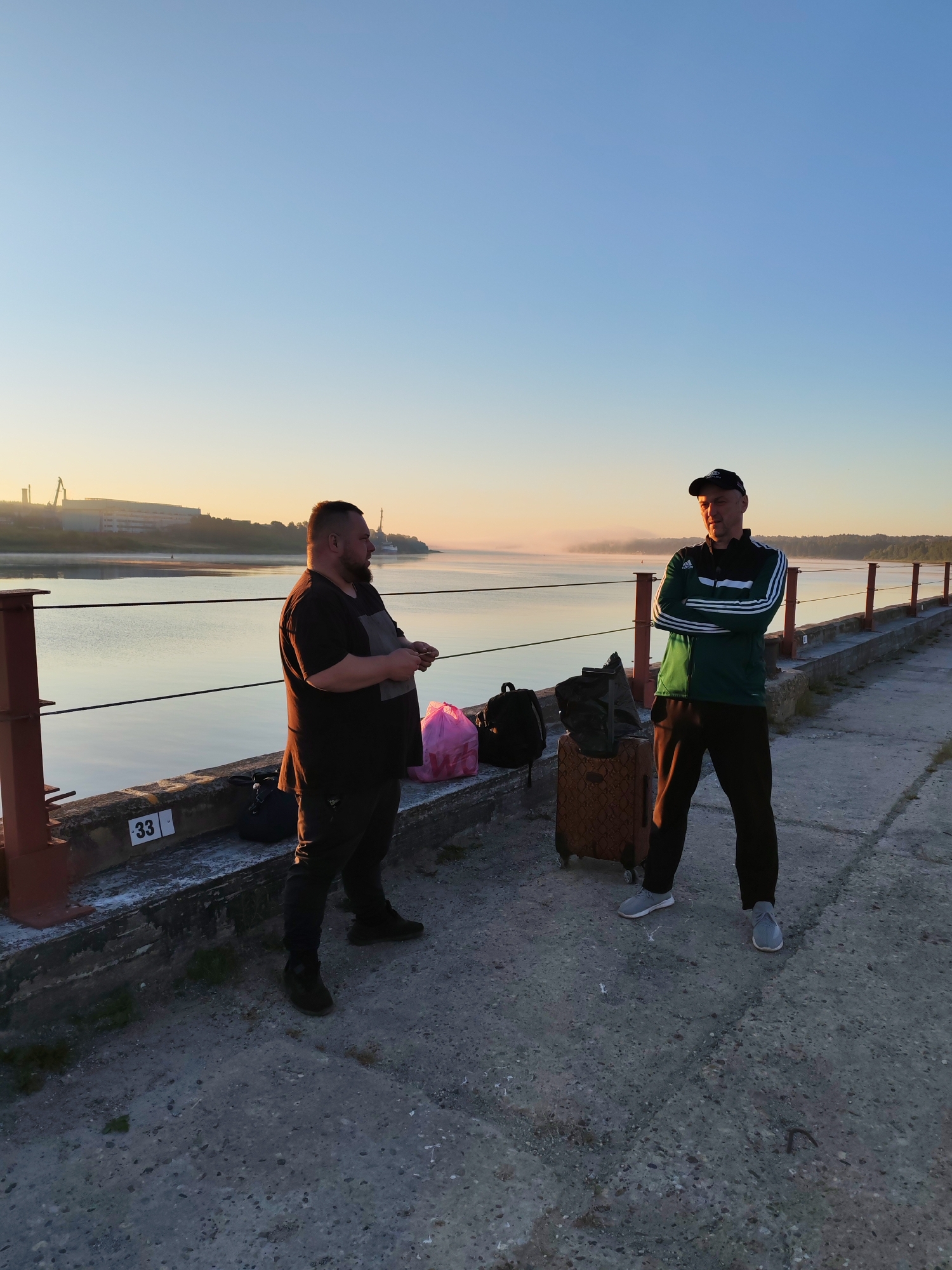 Two men stand on a concrete pier next to a body of water in Bol'shie Mkhi, Russia. The man on the left is heavier-set, wearing a dark t-shirt and dark pants. He is facing away from the camera, looking towards the water. Next to him are a pink bag and a black backpack. The man on the right is wearing a green and white tracksuit and a black baseball cap. He is facing the camera with his arms crossed. Beside him is a brown suitcase with wheels and a black backpack. In the background, there is a body of water with a thin layer of fog on its surface, and a distant industrial building. The sky is a clear blue with a hint of sunrise or sunset glow near the horizon. The pier has metal railings and concrete barriers. The pier has a number "33" marked on the barrier. The time of day appears to be either early morning or late evening, indicated by the soft lighting and the fog.