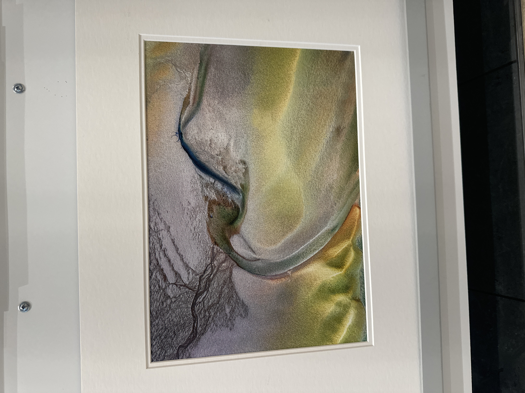 The image shows a framed abstract artwork, likely a photograph, hanging on a white wall. The artwork depicts an aerial view of a landscape characterized by flowing lines and a gradient of colors, ranging from dark gray and purple on the left to greens and yellows on the right. These colors suggest natural elements such as water, sand, and vegetation. The texture appears granular, resembling sandbanks or dried mud. The artwork is mounted on a thick, white mat and framed with a thin white border. Two small screws are visible on the wall above the frame, suggesting it is mounted directly to the wall. The bottom of the image shows a glimpse of a dark surface, possibly a shelf or countertop.
The question about the location in Kilkenny, Ireland, cannot be answered from the provided image. The image itself does not contain any visual cues or information that would identify a specific location within Kilkenny or any other city.