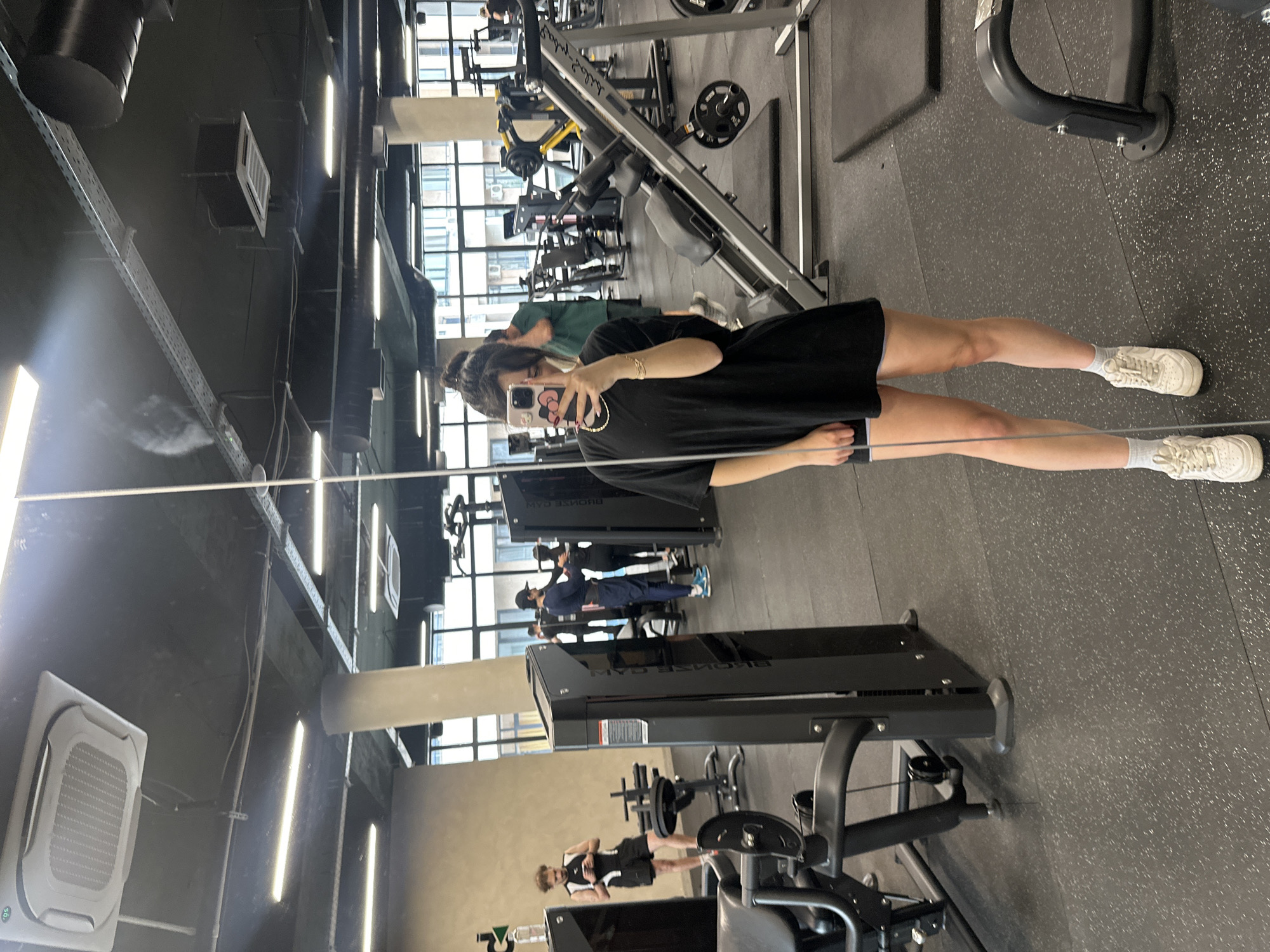 A person is taking a mirror selfie inside a gym. The individual is wearing a black t-shirt and shorts, with white sneakers and socks. They are holding a smartphone in their right hand, with a visible reflection in the mirror. Gym equipment is visible in the background. Other people are using the gym equipment. The location appears to be a gym with large windows. Based on the gym equipment and layout, it is unclear whether this is in the city of Yerevan, Armenia. Additional information or visual cues would be required to confirm the location.