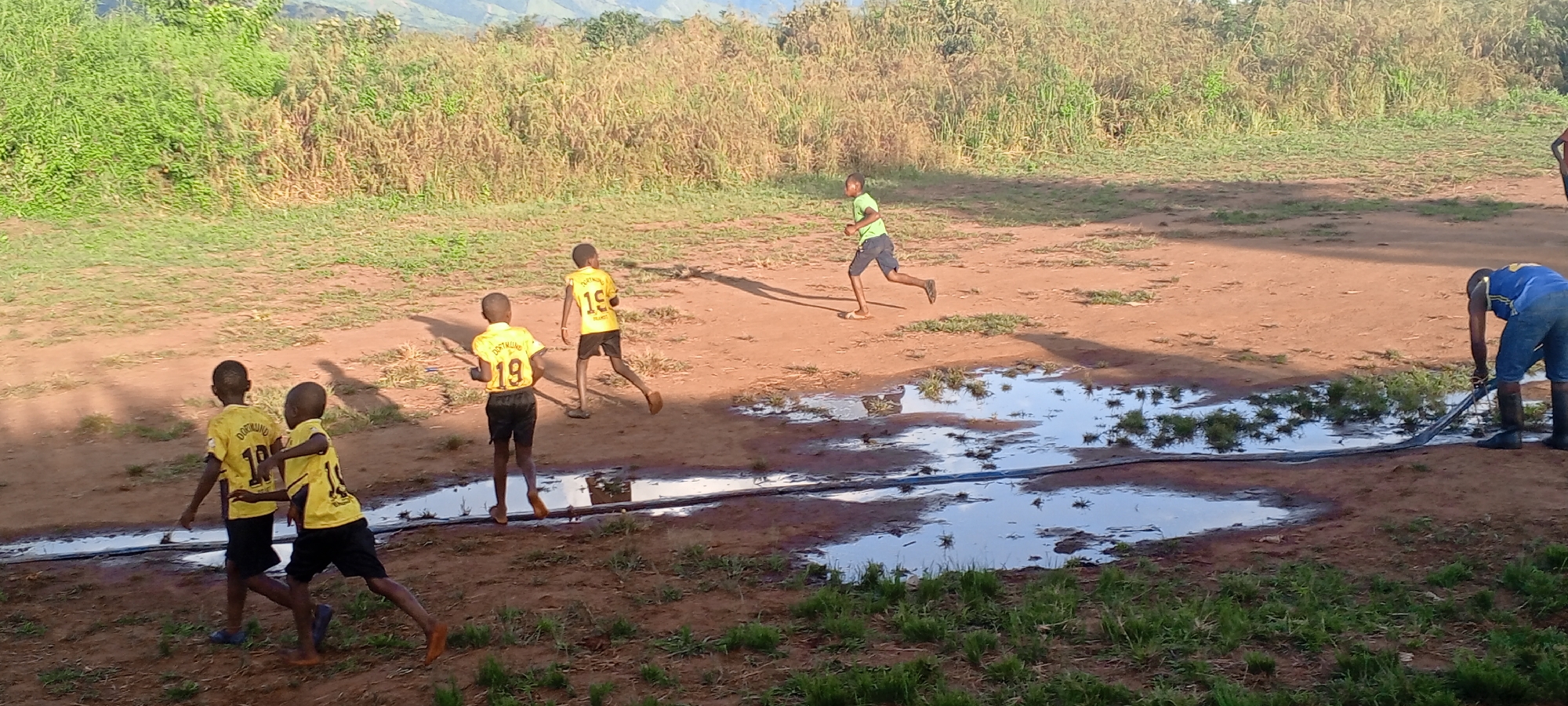 The image depicts a scene near Ngalula, Democratic Republic of the Congo. Several individuals are present. Four children wearing yellow Dortmund soccer jerseys with the number 19 are on the left side of the image. A fifth child wearing a green shirt is running near the center. A man wearing a blue shirt and boots is on the right, interacting with a long black hose near a large puddle. The ground appears to be dirt or mud, with puddles of water. There is vegetation in the background, including green grassy areas and taller shrubs. Distant hills are visible in the background.