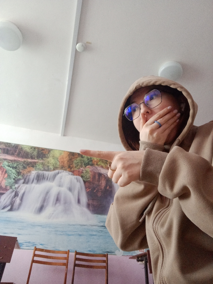 An individual is depicted indoors, viewed from a low angle, wearing a beige hooded sweatshirt with the hood raised and eyeglasses featuring blue lens reflections. The person's left hand covers their mouth, displaying a light blue ring on the ring finger. Their right arm is extended, with the index finger pointing towards the left; a gold-colored ring is visible on its ring finger.
The background features a large wall mural depicting a multi-tiered waterfall cascading into a blue body of water, flanked by autumnal foliage. Below the mural, two wooden chairs with slatted backs are visible. The lower wall section appears light pink. The white ceiling contains two circular, flush-mounted light fixtures and a small white cylindrical device, likely a smoke detector. A white beam or trim piece crosses the ceiling. The specific location within the city of Chaikovsky, Russia, cannot be determined from the visual information provided.