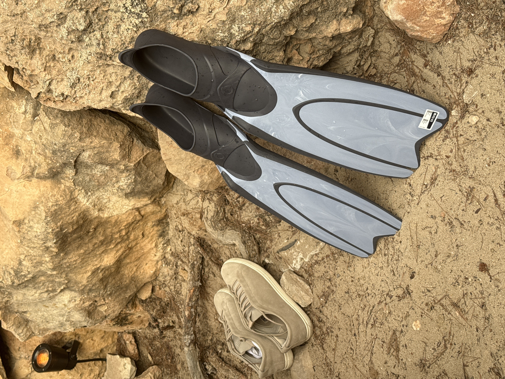 Two pairs of shoes and a pair of fins are resting on the sand in front of a rocky surface. A small spotlight is visible to the left of the scene, illuminating the rocks. The fins are black and gray, and the shoes are light brown. The scene appears to be outdoors, possibly near a beach or coastal area. The specific location of Sant Antoni de Portmany, Spain, cannot be determined from the visual information provided in the image.