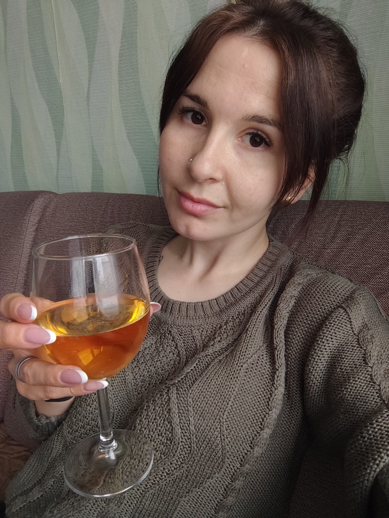 A young woman, likely in her 20s or early 30s, is depicted in a close-up, selfie-style photograph. She has dark hair, styled away from her face, and brown eyes. A small silver stud piercing is visible on her right nostril. She is wearing a chunky, cable-knit sweater in a muted olive-green or dark grey-green color. Her left hand, adorned with a ring on the ring finger and featuring French-tipped nails, holds a stemmed glass containing an amber-colored liquid. She looks directly at the camera with a neutral to subtly pleasant expression.
The setting appears to be indoors, likely a home, given the casual attire and background. She is seated on or leaning against a dark brown or taupe upholstered couch. Behind her, the wall is covered with a light green or teal wallpaper featuring a distinct wavy, striped pattern. The lighting suggests daytime, but no specific light source is apparent. There is no visible text in the image. The scene conveys a relaxed and casual moment in Фоки, Russia.