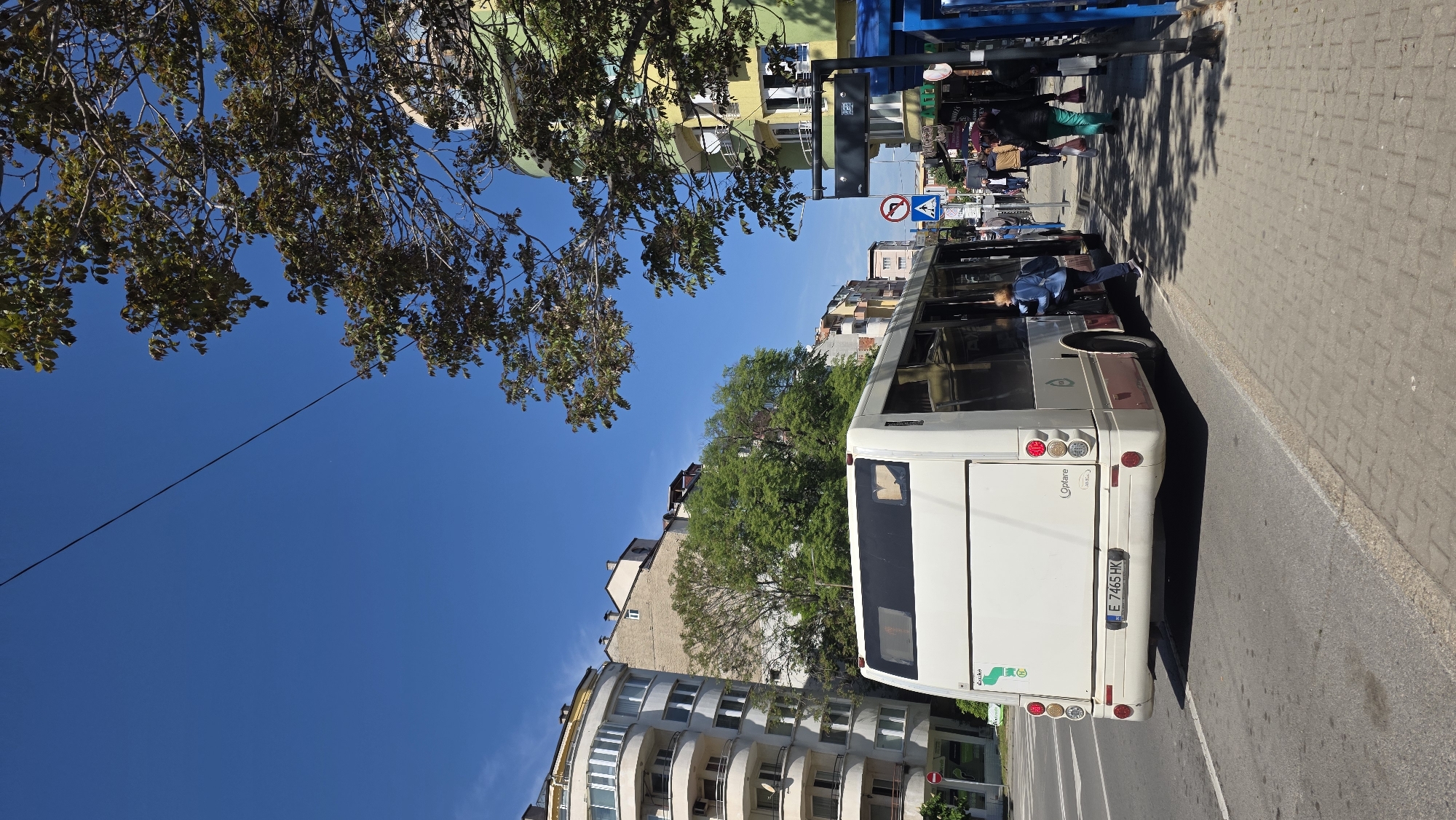 The image captures a street scene in Blagoevgrad, Bulgaria, featuring a white bus parked alongside a sidewalk. The bus, partially visible, has the license plate "E 7465 HK" and bears the "Optare" brand. A person wearing a blue shirt and dark pants is entering the bus. Pedestrians walk along the sidewalk. A "No U-Turn" sign is visible in the background. Buildings in various architectural styles line the street, with trees interspersed among them. The sky is clear and blue. A tree with full foliage is visible in the upper left corner of the frame. The perspective of the image is slightly tilted.