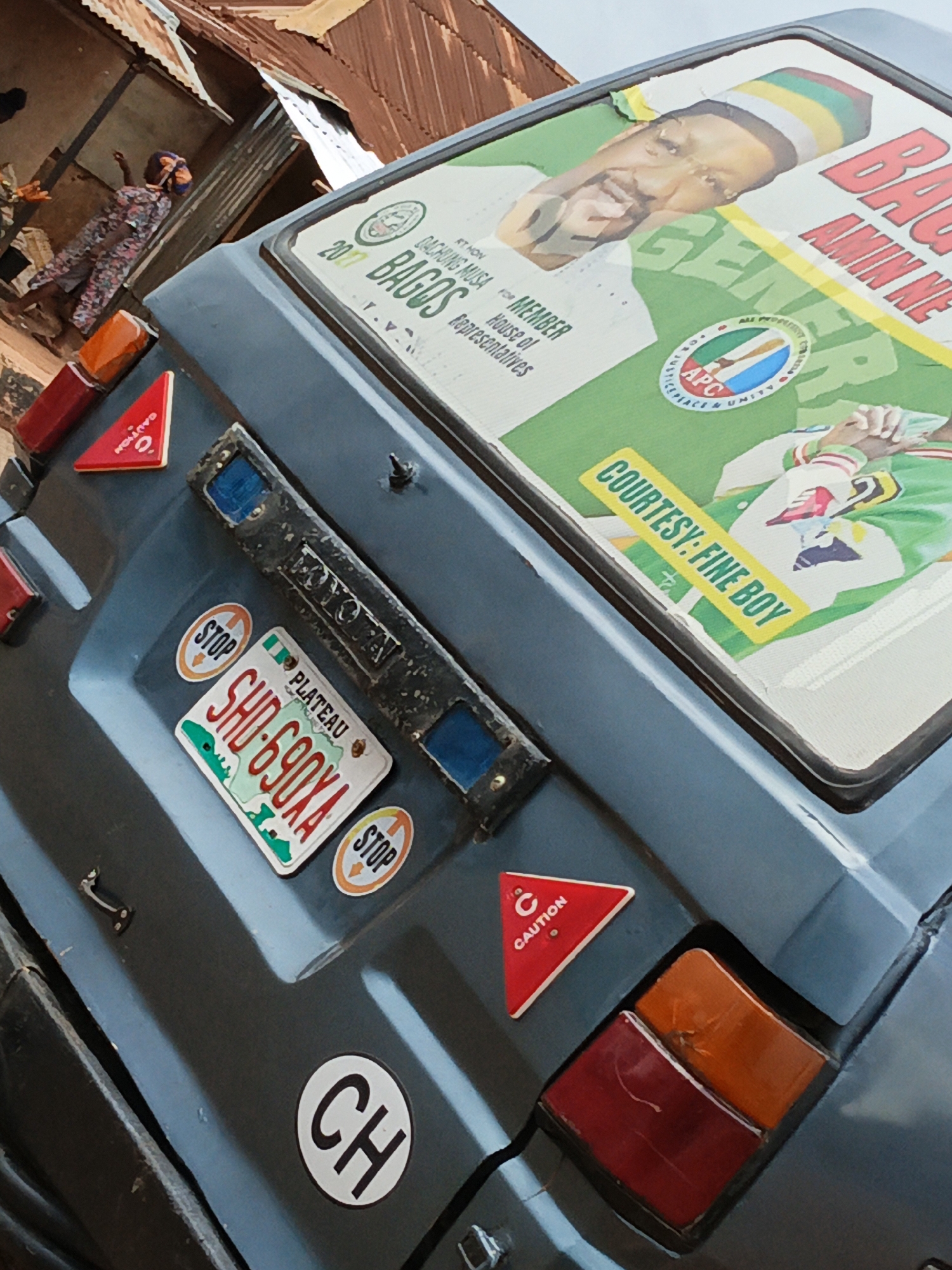 A side view of the back of a blue minivan is shown in this image. The minivan is covered in stickers and has a license plate that reads "SHD-690XA PLATEAU". There are two circular "STOP" stickers on the back of the van, as well as a triangular "CAUTION" sticker. A political poster is affixed to the rear window of the van, featuring the face of a man in a striped hat and the words "2017 BAGOS FOR MEMBER House of Representatives APC". The background of the image shows a corrugated metal roof of a building and a woman walking in what appears to be a market.