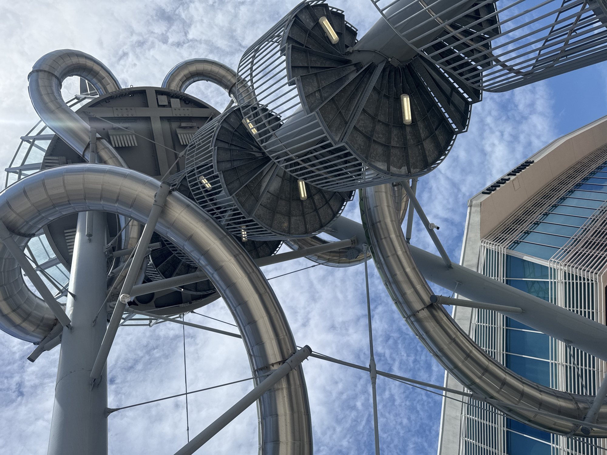 The image shows a low-angle view of the ArcelorMittal Orbit sculpture and slide in Queen Elizabeth Olympic Park, London, United Kingdom. The metallic structure features curved tubes and spiral staircases against a backdrop of a cloudy sky. A modern building with glass windows is visible in the lower left corner. There are no people or animals in the image. The scene is lit by daylight.