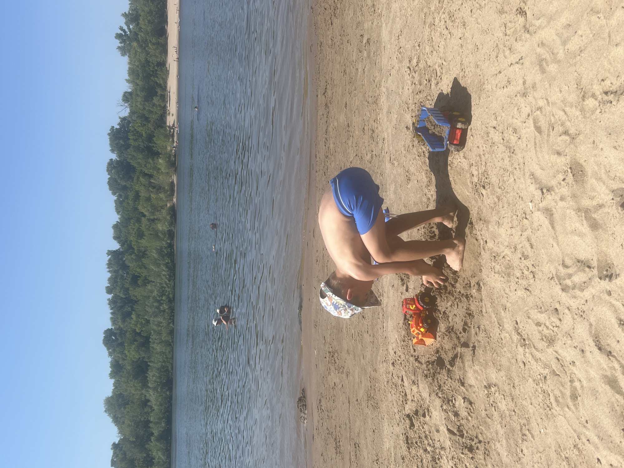 A young boy wearing a patterned cap and blue swim trunks is playing with toy construction vehicles on a sandy beach next to a body of water. He is crouched down, interacting with a red and orange toy tractor. To his right, a blue toy dump truck filled with sand is visible. In the background, people are swimming and wading in the water. Across the water, a densely wooded shoreline forms the horizon. The sky is clear and blue.