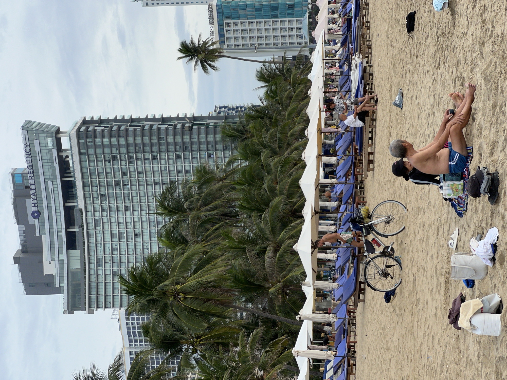 The image depicts a beach in Nha Trang, Vietnam, with a prominent Hyatt Regency hotel in the background. In the foreground, a sandy beach is covered with blue lounge chairs and white umbrellas. A bicycle with a basket filled with yellow fruit is parked on the sand. Two elderly men are seated on the sand, facing away from the camera, with one man wearing blue swim trunks and the other wearing a white shirt. Several bags and clothing items are scattered on the sand. In the mid-ground, a man with a beard is standing near the bicycle, holding a towel. The background shows a dense line of palm trees and several tall buildings, including the Hyatt Regency hotel, which is characterized by its modern architecture and glass facade. The sky is overcast.