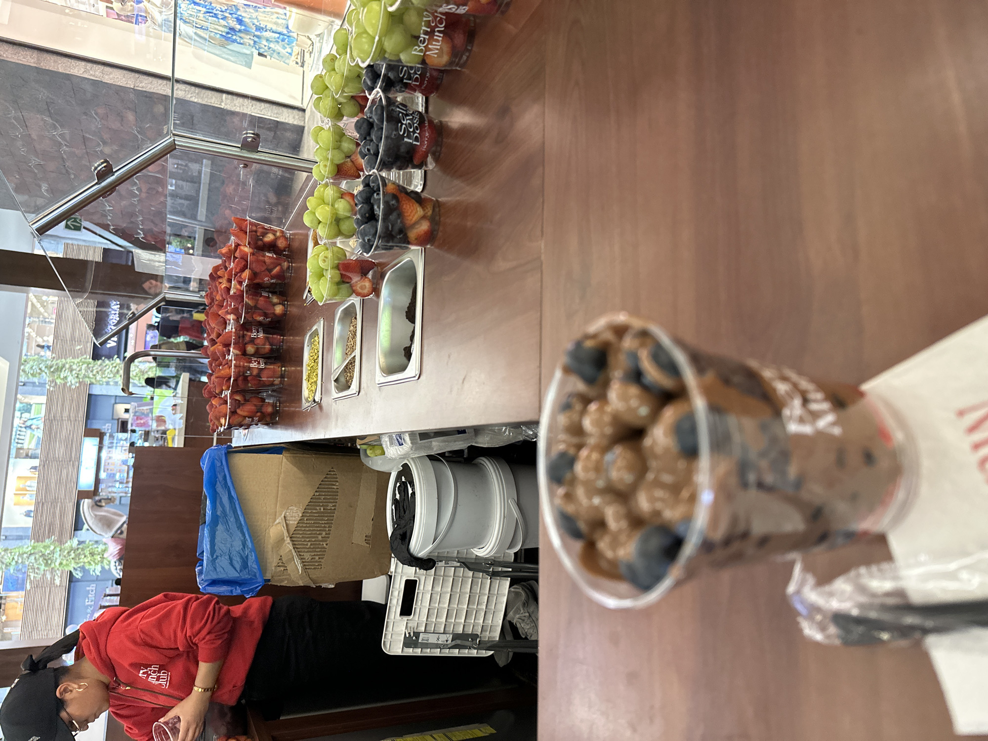 This image depicts an indoor fruit and dessert stall located within a shopping mall in Puebla, Mexico. In the foreground, a blurred clear plastic cup containing small, round, brown-colored items (likely chocolate-covered fruit or dough) mixed with blueberries rests on a white napkin.
The midground features a wooden service counter displaying numerous clear plastic cups filled with prepared fresh fruit, predominantly cut strawberries, green grapes, and blueberries. Several rectangular metal inserts are embedded in the counter, holding toppings such as chopped pistachios, a brown granular substance (likely granola), and dark crumbs (possibly chocolate). A clear glass protective barrier with chrome fittings separates the serving area from the customer space.
Behind the counter, an adult female employee is visible on the left. She wears a red hoodie with "Berry Munch Club" branding, a black baseball cap, eyeglasses, and a gold hoop earring. She is holding a clear plastic cup in her left hand, appearing to prepare or arrange its contents. Behind her, a large cardboard box lined with a blue plastic bag serves as a waste receptacle. Two white plastic buckets are stacked on the floor next to a white plastic crate, and black gloves are placed on the top bucket. In the background, beyond the stall, glimpses of other retail storefronts, including "Abercrombie & Fitch" and "Victoria's Secret," are visible within the multi-level mall environment.