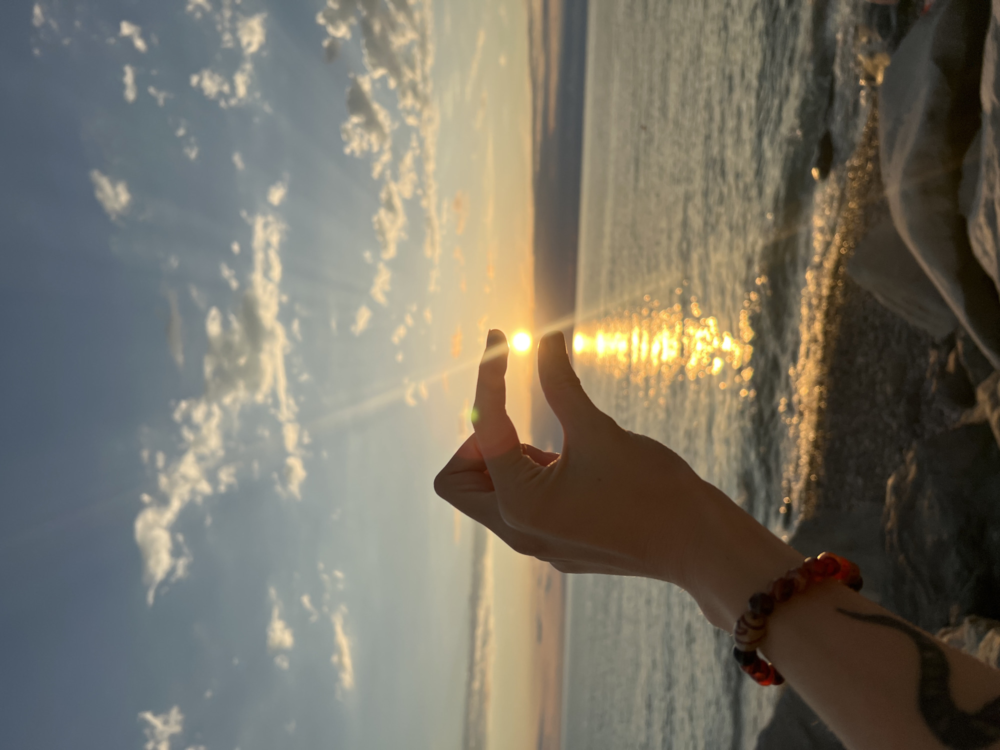 A hand is held up against a sunset over the sea. The sun is partially obscured by the fingers, creating a lens effect. The sky is a gradient of orange and blue, with scattered clouds. The sea reflects the sunlight, creating a shimmering path. The foreground shows a rocky shoreline and the arm of the person holding the sun, adorned with a beaded bracelet. The location is the beach in Batumi, Georgia.