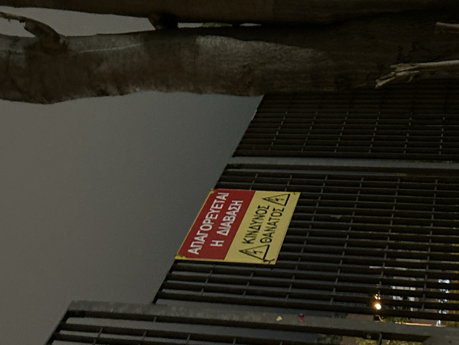 The image captures a dark outdoor scene in Kallithea, Greece, likely at night or dusk, showing a metal grate structure and a large tree limb. The camera perspective is rotated, making the tree limb appear at the top and the metal grate angling down to the right. The grate, possibly part of a bridge or platform, is made of dark, evenly spaced metal bars. Below the grate, the area is dark, with some distant artificial lights visible, suggesting a drop or open space beneath.
Attached to the metal grate is a prominent warning sign with red and yellow sections. The red section, on the left, contains the Greek text "ΑΠΑΓΟΡΕΥΕΤΑΙ Η ΔΙΑΒΑΣΗ," which translates to "PASSAGE IS FORBIDDEN" or "NO PASSING." The yellow section, on the right, features a black lightning bolt symbol indicating electrical hazard, followed by the Greek text "ΚΙΝΔΥΝΟΣ ΘΑΝΑΤΟΣ," translating to "DANGER DEATH."
No people or direct activity are visible, but the warning sign strongly implies a dangerous area where entry or passage is prohibited, possibly due to electrical hazards or a fall risk. The overall mood conveyed is one of caution and potential danger.