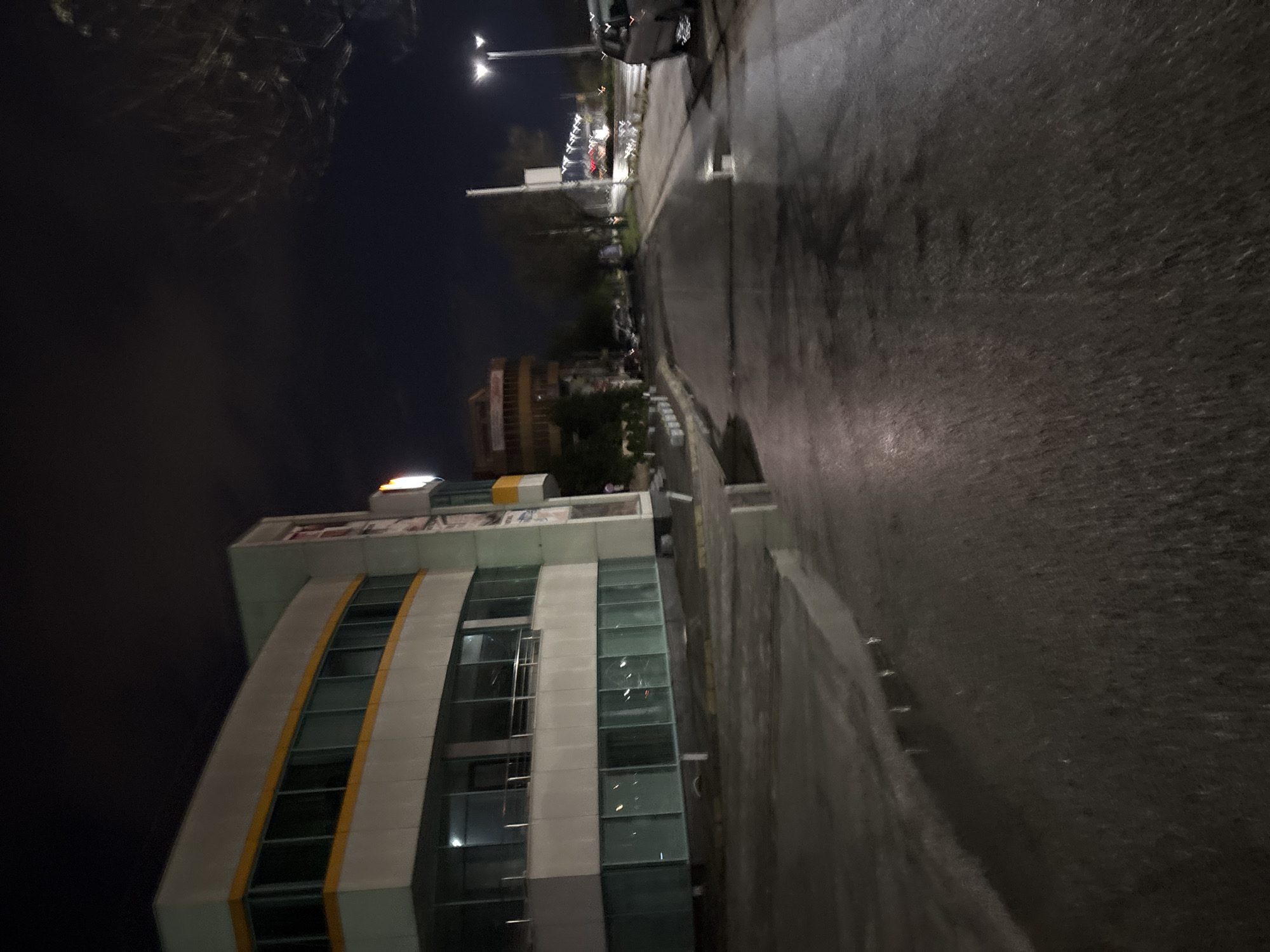 The image shows an urban night scene in Plovdiv, Bulgaria. A modern building with curved architectural lines and glass windows is on the left, illuminated by internal lights. The street beside the building is dark and appears wet, possibly from rain. Streetlights illuminate the street in the distance. There is a car parked along the street further down. The sky is dark, with minimal cloud cover. The image is taken from a low angle, causing a distorted perspective.