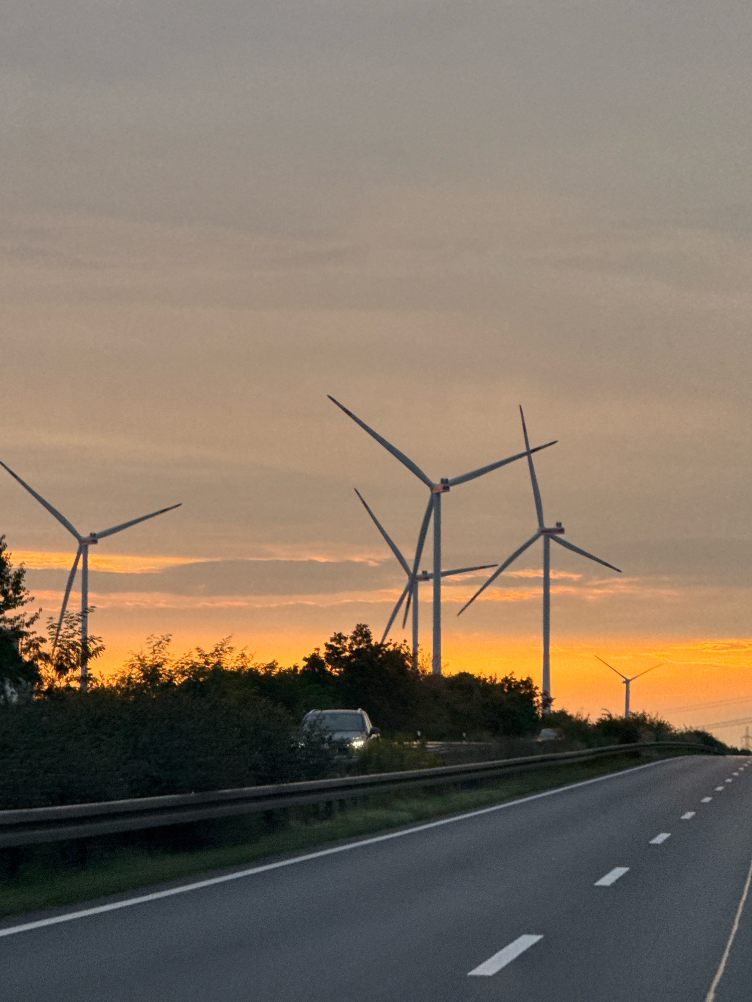 The image depicts a highway with wind turbines in the background at sunset. The sky is a gradient of orange and grey hues. Several wind turbines are visible, with their blades extended. In the foreground, the highway is paved with a guardrail on the left and dotted white lines indicating lanes. A dark-colored car is visible on the side of the road behind the guardrail. The overall scene suggests a rural or semi-rural setting near Salzgitter, Germany, given the presence of wind turbines.