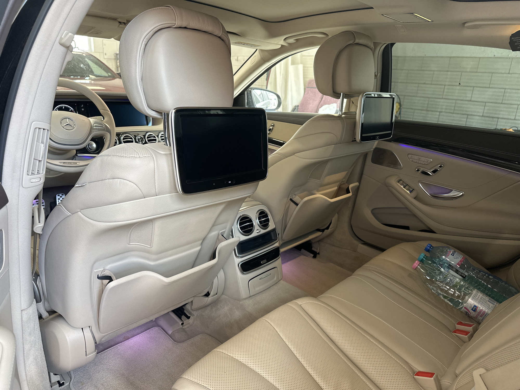 The image shows the interior of a luxury car, likely a Mercedes-Benz S-Class, viewed from the rear passenger seats looking forward. The interior features cream-colored leather seats with perforation, and ambient lighting is visible, casting a purple hue on the floor mats and lower parts of the car.
In the foreground, the right rear passenger seat is visible, with two plastic water bottles placed on the seat cushion. The bottles are labeled in Cyrillic script, with one appearing to be "DEVIN" brand mineral water. The seat belts are buckled.
The back of the front passenger seat is visible to the left, with a built-in entertainment screen and storage pockets. The driver's seat and steering wheel are partially visible through the gap between the front seats. The steering wheel is also cream-colored leather with a Mercedes-Benz emblem.
The rear of the driver's seat is to the left, also featuring a storage pocket. Between the front seats, the center console with air vents, controls, and a small storage compartment with a pull-down door is visible.
In the background, the left rear seat is visible, and another entertainment screen is mounted on the back of the front driver's seat. The car's panoramic roof is visible at the top of the image.
The environment outside the car, visible through the windows, suggests an indoor setting, possibly a car wash or a garage. The walls appear to be made of brick or concrete blocks. In the far background, through the windshield, another car, possibly a dark red sedan, is partially visible.
While the image itself does not contain explicit indicators of the city of Burgas, Bulgaria, the presence of Cyrillic script on the water bottles, a common characteristic of products sold in Bulgaria, strongly suggests that the image was taken in Burgas, Bulgaria. The car appears to be stationary.
