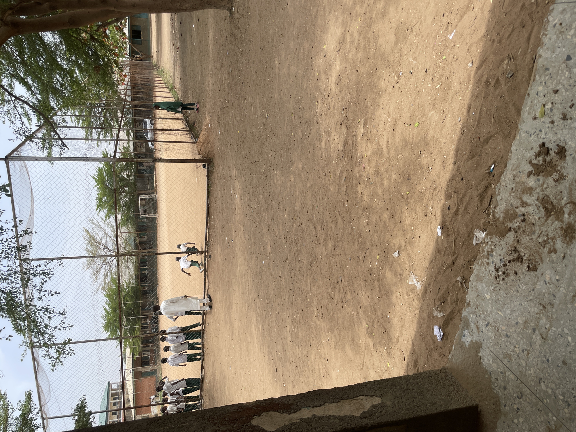 The image captures an outdoor scene at what appears to be a school or community ground in Unguwar Rimi, Nigeria.
In the foreground, a dusty, light-brown earth ground covers most of the area, transitioning to a rough concrete or paved edge on the far right. Small pieces of litter, including paper and plastic, are scattered across the dirt.
A metal chain-link fence runs across the midground, separating the immediate foreground from an active area beyond. Behind this fence, on the left side, a group of approximately 8-10 young individuals, likely students, are lined up. They are wearing white shirts and dark trousers or skirts, suggesting school uniforms. An adult or older youth, dressed in a long white thobe and dark pants, stands near this group, seemingly supervising or addressing them.
Further into the fenced area, to the right, two other individuals wearing white shirts and dark shorts are visible, one in motion, possibly running or playing. A small soccer goal is situated near the center-left within the fenced space, indicating a sports or play area. At the far right end of the fenced enclosure, a lone person in a green outfit stands, observing the scene.
In the background, beyond the fenced area, trees with green foliage are prominent, and parts of buildings, possibly school structures, are visible. A white car can be faintly seen through the fence in the distance.
The activity appears to be a gathering or sports practice involving the students, possibly soccer, given the goal and the individuals in athletic attire. The bright sunlight and distinct shadows suggest it is daytime, likely mid-morning or afternoon, under clear weather conditions. There is no visible text in the image.