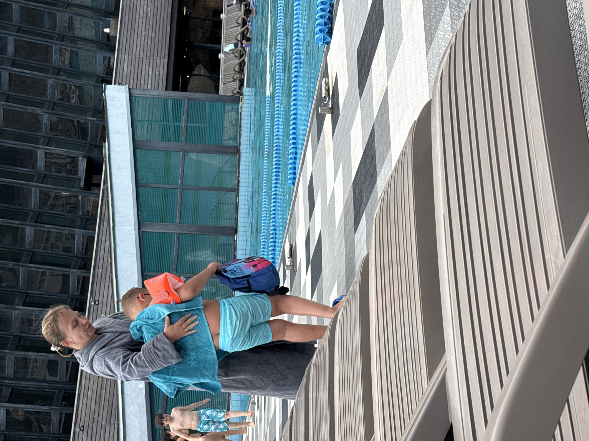 This media file shows a scene at an outdoor swimming pool, likely in Yalta, Ukraine. The image is oriented sideways. In the foreground, a woman wearing a grey robe is holding a young child. The child is wearing light blue swim trunks and an orange flotation device on their upper body. The child is also wrapped in a blue towel and carrying a backpack with a colorful design.
To the left of the woman and child, there is a tall building with a dark, reflective glass facade, suggesting it's a modern structure. Adjacent to this building is a section with wooden paneling. A bright turquoise pool with blue lane markers is visible further back. The pool area is bordered by a patterned surface with a checkered design of grey and white squares, and a light-colored slatted fence or railing.
In the background, other people are partially visible near the pool. The lighting suggests it is daytime, likely sunny, given the reflections on the building and the brightness of the scene. The overall atmosphere appears to be casual and relaxed, consistent with a day at a swimming pool. There is no visible text in the primary focus of the image, though the flotation device may have text that is not legible.