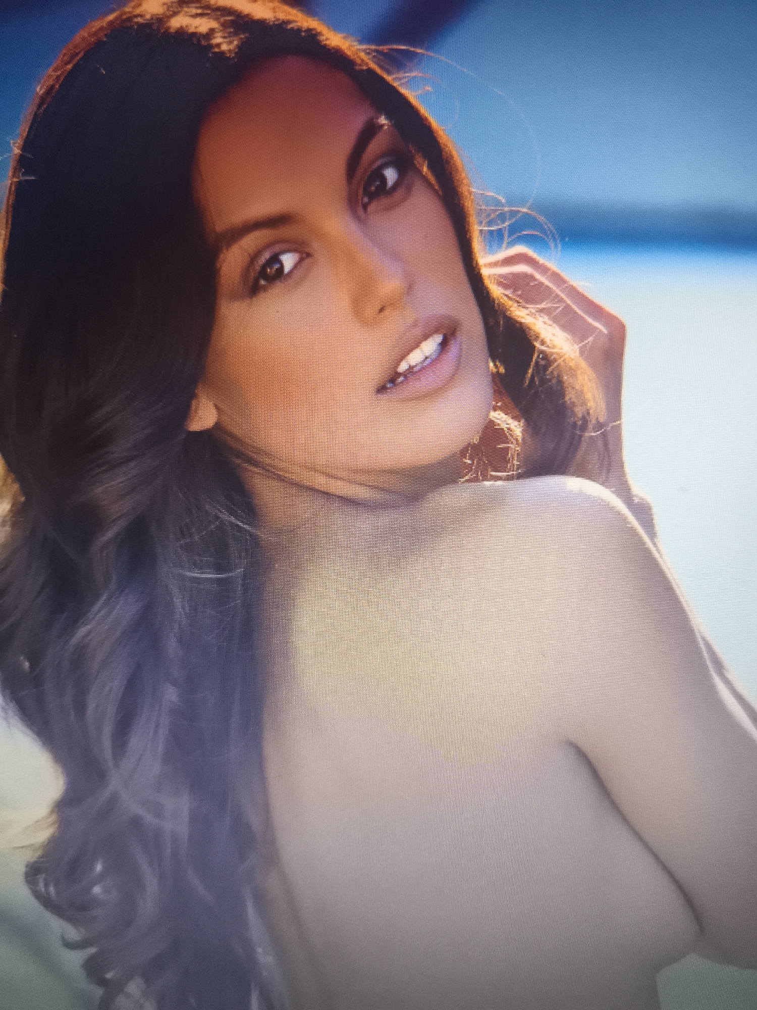 A woman with long, dark brown hair is facing the camera and looking over her left shoulder. She has light skin and her lips are slightly parted. Her right hand is near her head, with her fingers lightly touching her hair. The background consists of a blue and white gradient, possibly water and sky. The image does not appear to be in Nowy Tomyśl, Poland.