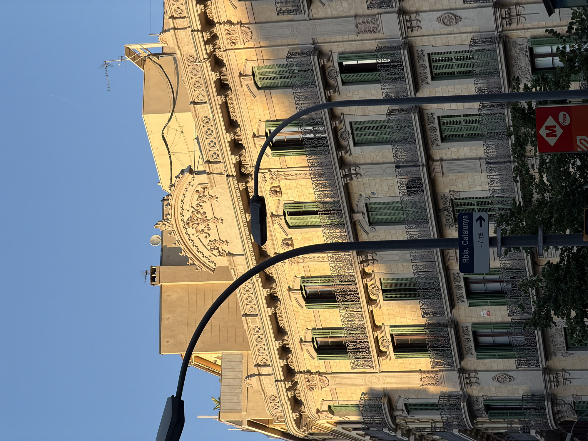 The photograph captures a facade of a building in Barcelona, Spain, likely on a street with historical architecture. The building is constructed of light-colored stone blocks and features multiple stories with ornate balconies. The balconies have black wrought-iron railings and are adorned with decorative elements. Numerous windows with green shutters are visible on each floor. A street sign indicating "Rbla. Catalunya" and an arrow pointing to the right suggests the location is La Rambla de Catalunya. A streetlamp is prominently featured in the foreground, its curved arm extending across the image. Below the street sign, a red sign with a white "M" inside a diamond, likely representing the Metro transport system, is visible. The sky is a clear blue, and the warm sunlight casts shadows on the building, indicating it is either early morning or late afternoon. Lush green trees are partially visible at the bottom of the frame, suggesting a well-treed urban environment. The architectural style points to the late 19th or early 20th century, characteristic of Barcelona's Eixample district.