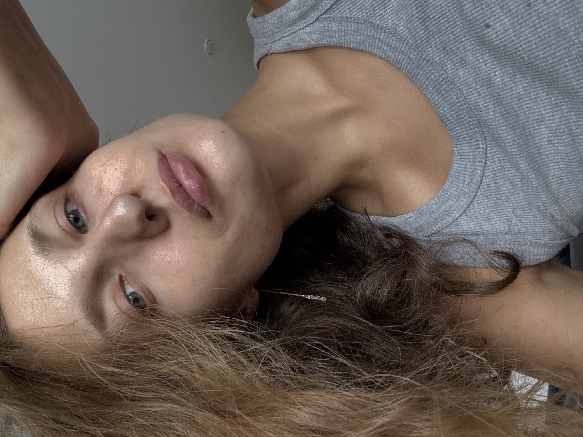 A medium shot shows a fair-skinned young woman with light brown hair, parted on her left, and blue eyes. She is looking towards the camera with a neutral expression. Her right hand is resting on top of her head. She is wearing a grey ribbed tank top. A delicate gold earring with three small clear stones hangs from her left ear. The background is a plain grey wall. The image was taken indoors.