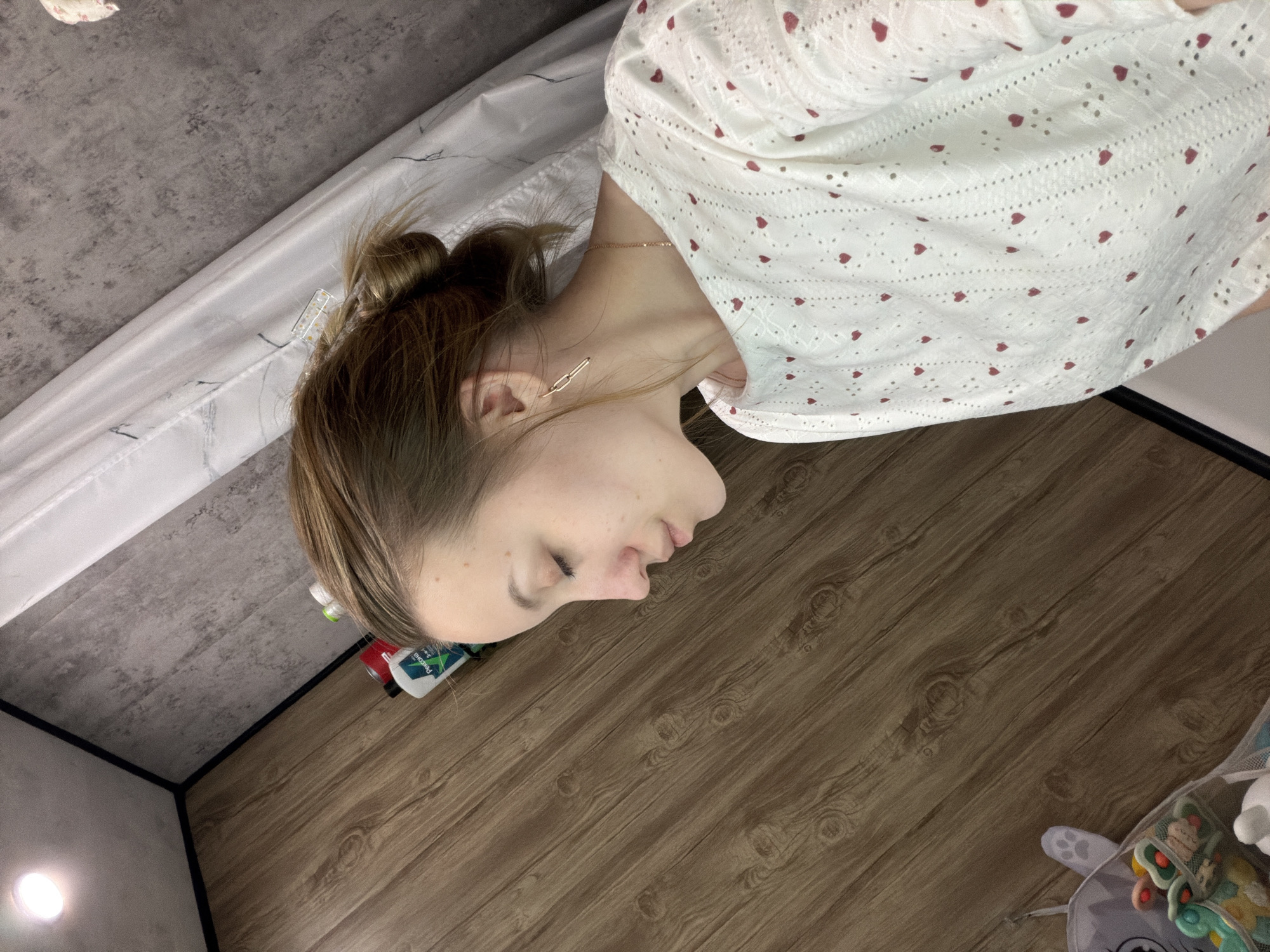 A woman with light skin and light brown hair in a bun is lying on a wood-patterned floor. She is wearing a white top with small red heart patterns and gold earrings and a necklace. Her eyes are closed and her face is in profile. The floor is brown with a wood grain pattern. In the background, there is a gray wall and a white surface. Near the woman's head, there are bottles of cleaning products and a red can. In the bottom right corner, there is a baby play mat with toys. The location is potentially Reshetnikovo, Russia, but this cannot be verified solely from the image.