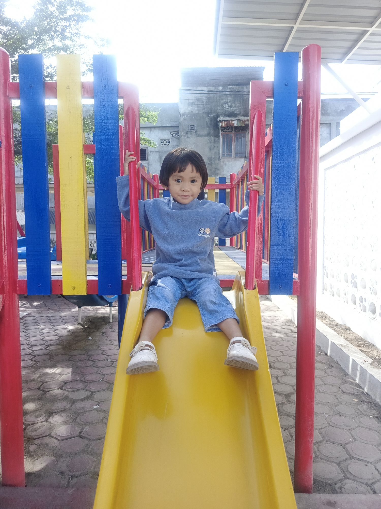 The image presents a young child positioned on a yellow slide within an outdoor playground in Pantegajah, Indonesia. The child, who appears to be between three and five years old, has short, straight, dark hair and dark eyes, and is looking directly at the camera with a neutral expression. The child is dressed in a light blue long-sleeved fleece top with a small embroidered emblem on the left chest, light blue denim shorts, and white closed-toe sandals with straps.
Both of the child's hands are gripping red vertical support bars on either side of the yellow slide, which is part of a larger playground structure. This structure features prominent elements of red, blue, and yellow painted wood or metal posts and railings. The ground surface beneath and around the playground consists of reddish-brown and grey hexagonal paving stones, showing some patches of dirt and wear.
In the background, an older, grey concrete building with a dark roof and visible barred windows is present on the left side. To the right, a bright white wall with patterned circular cutouts extends along the edge of the play area. Above the playground structure, parts of green tree foliage are visible against a bright, overexposed white sky, indicating strong overhead sunlight. A light-colored corrugated roof structure is also visible high on the right side of the background.