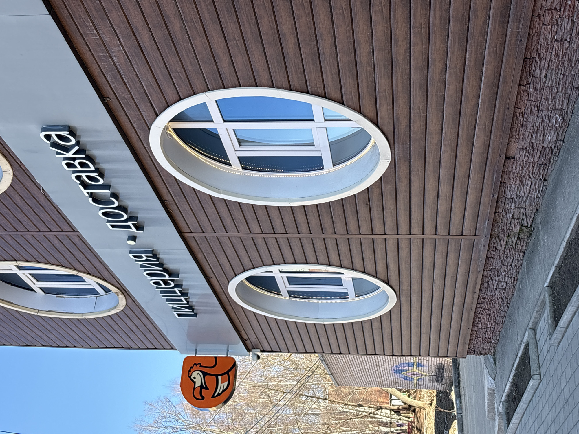 This daytime image showcases the facade of a building in Votkinsk, Russia. The building's exterior is predominantly covered with dark brown, vertically oriented wood-like paneling. A grey horizontal band runs along the upper part of the facade, featuring prominent white, three-dimensional letters with black outlines. This signage spells out "Пиццерия" (Pizzeria) and "Доставка" (Delivery).
Several distinctive oval-shaped windows are integrated into the brown paneling. Two such windows are clearly visible, with a third partially seen on the far left. These windows have white frames, multiple panes, and reflect the bright blue sky. Each oval window also appears to have a strip of small lights, possibly LEDs, running along its inner rim.
Below the main signage, to the left, an orange, shield-shaped sign hangs, depicting a stylized logo of a chicken or rooster head. To the right of the wood paneling, a section of the wall features a rough, textured brown material, possibly decorative stone or brick. The scene includes a sidewalk and a curb in the foreground. In the background, bare trees are visible against a clear blue sky, suggesting a bright day in cooler weather. No individuals are present in the image.