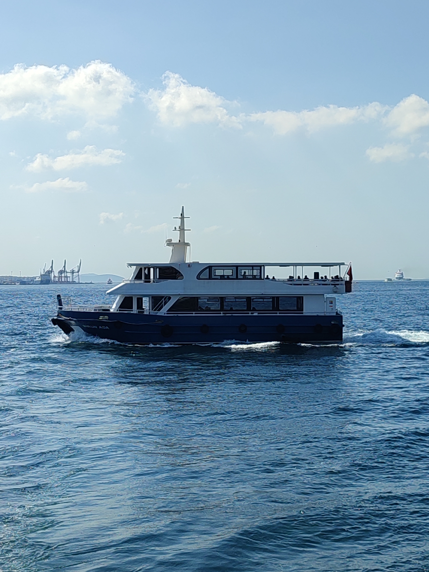 A ferry named "BEZIRGAN ADA" sails on the Bosphorus Strait, with the city of Beyoğlu, Turkey, visible in the background. The ferry, painted navy blue and white, has two levels and passengers can be seen on the upper deck. In the distance, industrial cranes suggest the presence of a port. The water is choppy, with white foam trailing the ferry. The sky is blue with scattered white clouds.