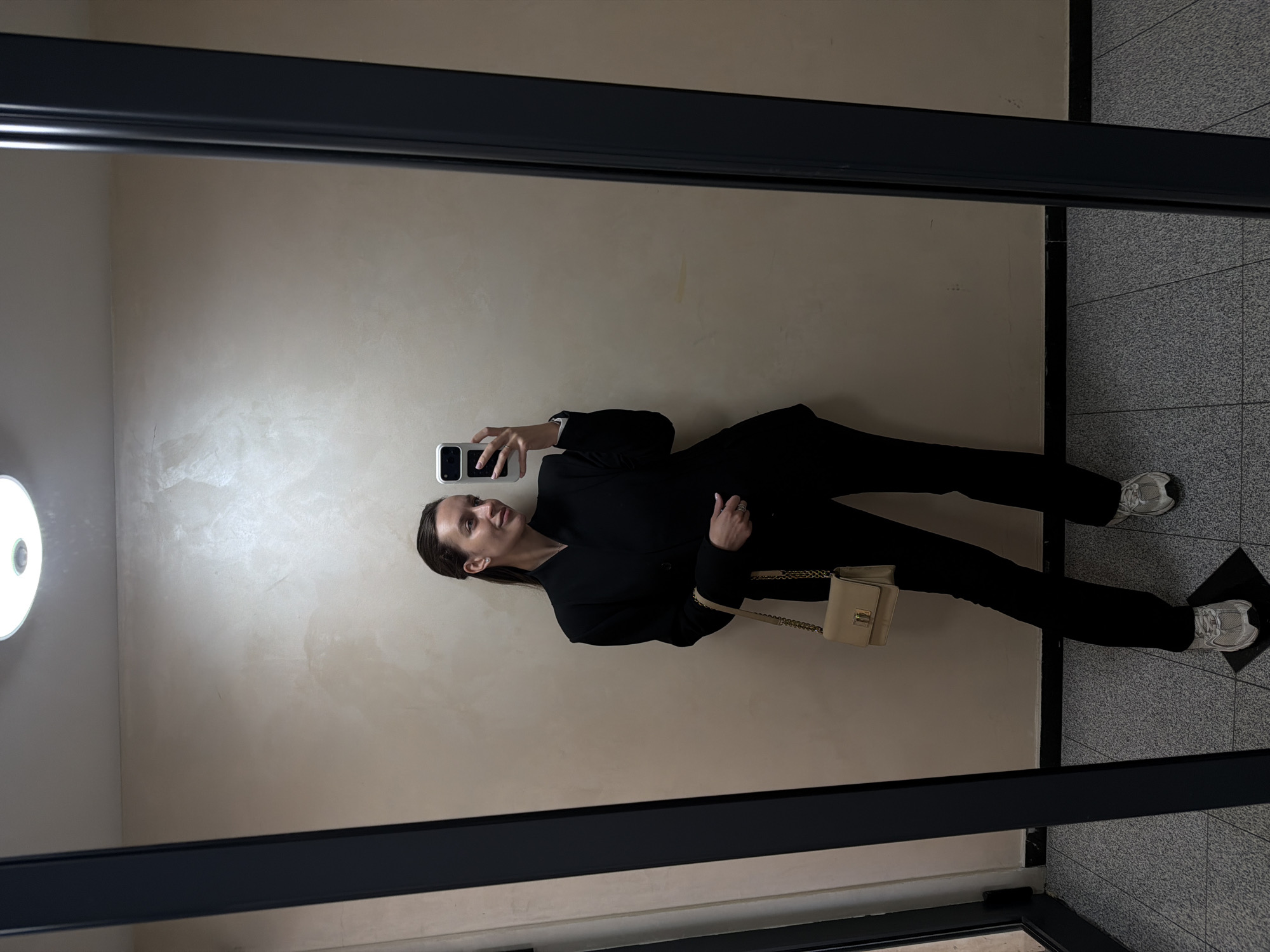 A woman is taking a selfie in a mirror. She is wearing a black blazer, black pants, and white sneakers. She is also carrying a light-colored handbag. The background consists of a beige wall on one side and tiled flooring on the other. The location is likely within the Vozdovac Urban Municipality, Serbia.