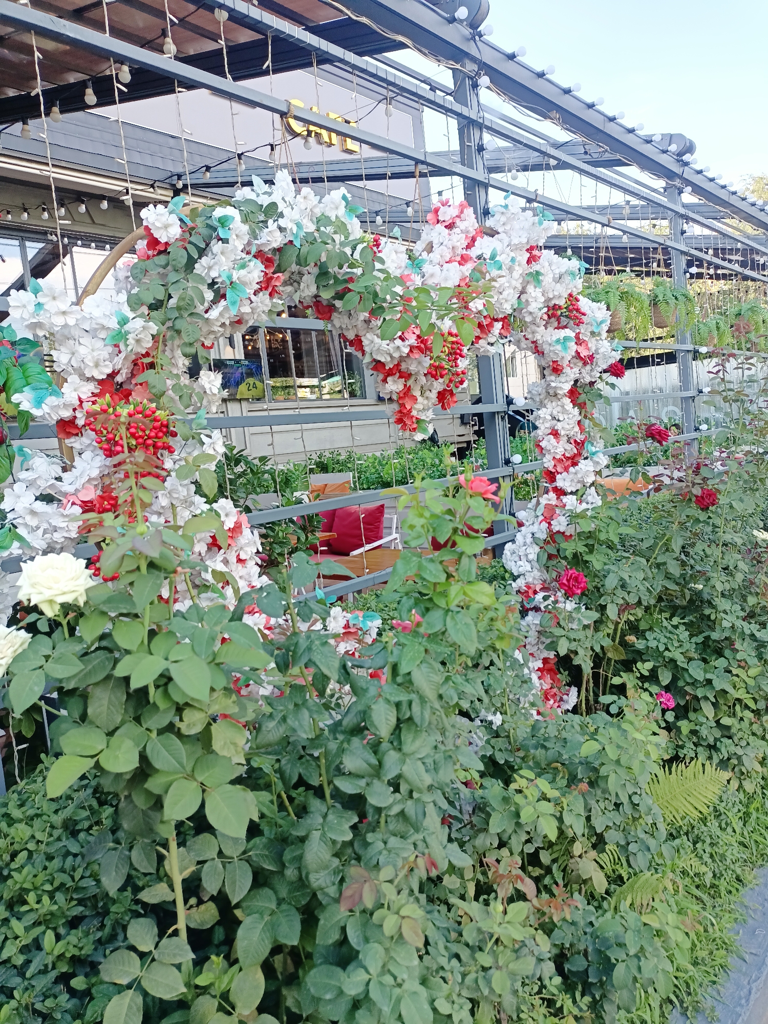 A horizontal, eye-level shot captures an outdoor seating area at what appears to be a cafe or restaurant in Almaty, Kazakhstan. The foreground is dominated by lush green rose bushes, with a prominent white rose bloom on the left. Behind the bushes, a large, heart-shaped archway constructed from artificial white and red flowers, along with red berries and small green and blue butterflies, is suspended from a metal pergola. This archway is adorned with string lights. Beyond the arch, an outdoor seating area is visible, featuring wooden tables and chairs with bright red cushions. A glimpse into the interior of the building reveals a sign with the number "24." The overall scene is vibrant and decorative, suggesting a welcoming atmosphere for patrons.