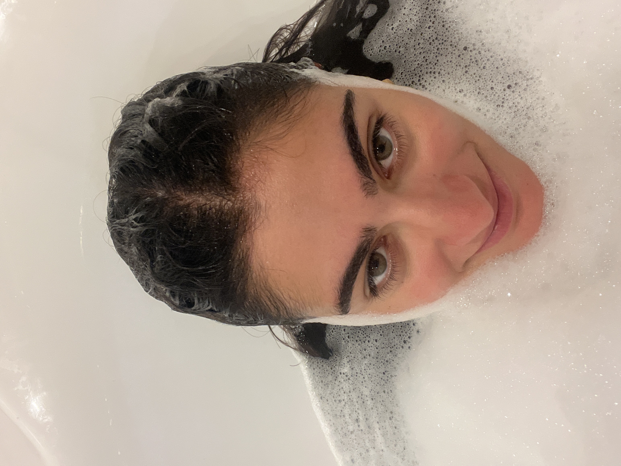 The image is a close-up, overhead shot of a young woman's face as she lies in a bathtub filled with white foam. Her dark, wet hair is slicked back, and some strands are falling forward. Her eyes are open and looking directly at the camera, with a hint of a smile on her lips. Her face is partially obscured by the foam, which reaches up to her chin and covers her neck. The bathtub is white and appears to be made of porcelain or acrylic. The lighting is bright, suggesting an indoor setting. The image does not contain any elements that would indicate a specific location in Almancil, Portugal.