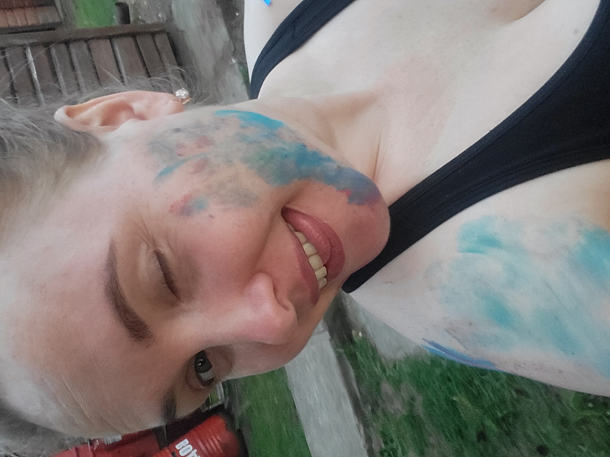 A fair-skinned woman with graying hair pulled back is winking and smiling at the camera. Her face and chest are smeared with blue and red paint, suggesting a recent activity like a Holi festival. She is wearing a black tank top. The background is blurred, showing a wooden fence and green grass, possibly indicating an outdoor setting in Prikamskii, Russia.