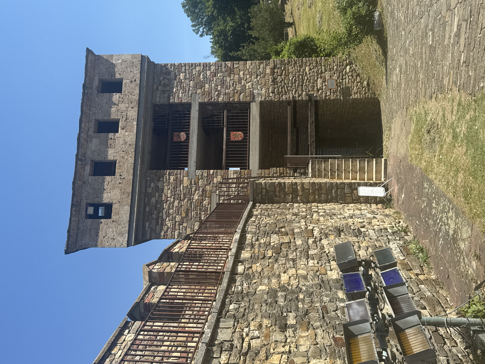 The image depicts a reconstructed medieval tower and wall, likely part of a historical site or attraction. The stonework is rough-hewn and appears aged. The tower has multiple levels with visible windows and balconies. Two banners with heraldic symbols are displayed on the balconies. A staircase leads up to one of the levels. In the foreground, a series of industrial spotlights are mounted on a pole against the stone wall. The ground is paved with stones in some areas and covered with grass in others. A small white sign on an easel stands near the staircase. The background shows a wooded area and a clear blue sky. This structure is identified as a reconstruction of a Roman watchtower, specifically part of the Roman fortress of
"Nicopolis ad Istrum", located in the vicinity of Veliko Tarnovo, Bulgaria. There are no people visible in the image.