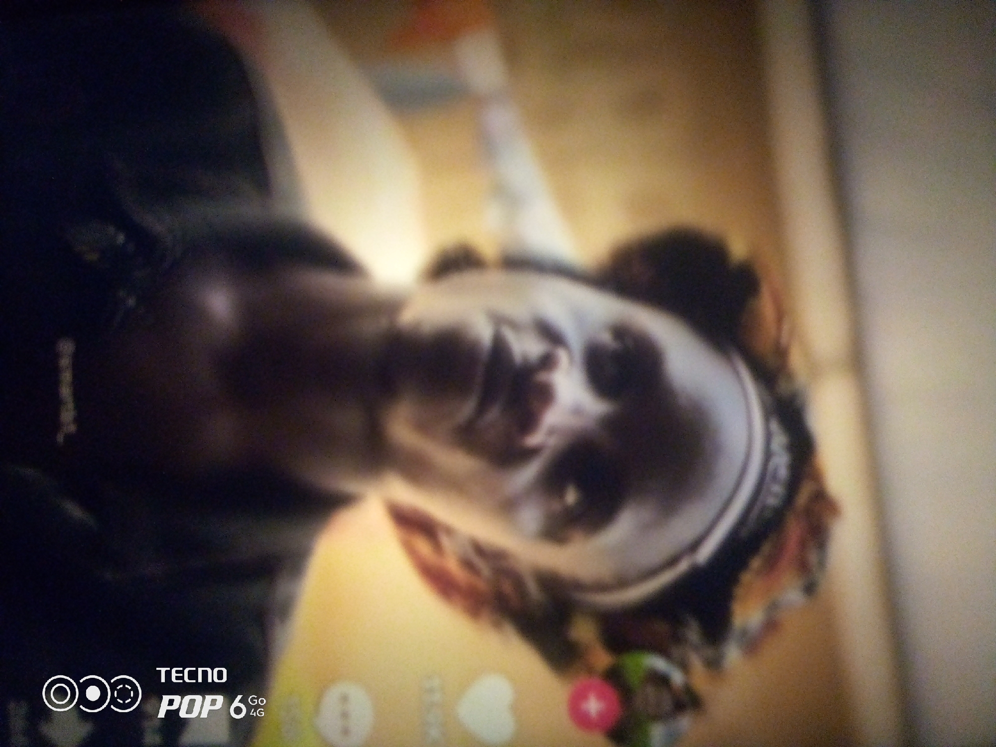 The image is a close-up, tilted, and somewhat blurry photograph of a person's face, which appears to be of African descent. The lighting suggests an indoor environment, possibly a living space, with a warm, yellowish light source. The person's head is tilted back, and their eyes are partially closed, giving the impression of relaxation or possibly sleep. In the lower left corner, there is a watermark that reads "TECNO POP 6 4G", indicating the device used to capture the image. The background is out of focus but suggests a wall or furniture. There are no explicit indicators of the exact location within Howayi, Nigeria, beyond the watermark suggesting a Nigerian context.