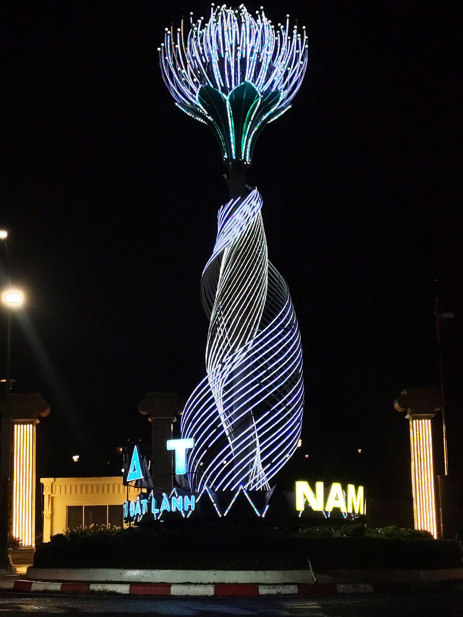 The image captures a nighttime view of a lighted monument in Da Lat, Vietnam. The monument features a base surrounded by shrubbery and a red-and-white painted border. Above the base is a structure with spiraling illuminated lines, white and blue in color. The top of the monument is designed as a flower-like form, with lighted elements extending upwards. The words "ĐẠT LẠNH" and "NAM" are illuminated in neon, visible at the base of the monument. A pair of illuminated pillars flank the monument, and a building is visible behind them. A street light is positioned to the left of the monument. The background is dark.