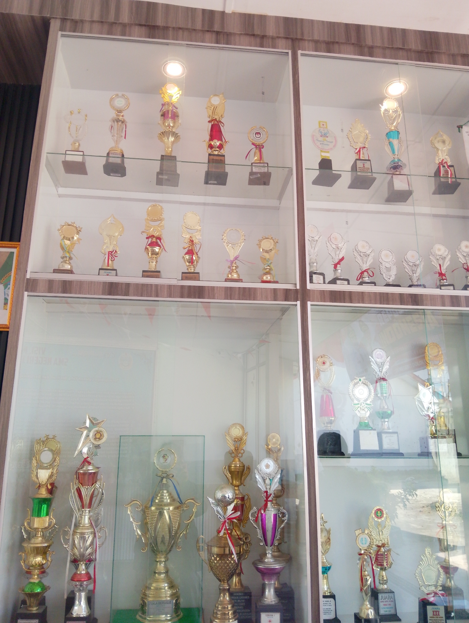 The image shows a wall of glass display cases filled with numerous trophies. The trophies are of various sizes and designs, mostly gold and silver, with some featuring ribbons and other decorative elements. They are arranged on shelves within the display cases, with some cases having internal lighting that illuminates the trophies. The background of the display cases appears to be a neutral, light color. The overall scene suggests a collection of awards, likely from a school, club, or competition. The context of Serbelawan, Indonesia, indicates that this is likely a public building or institution in that city. There are no people visible in the image.