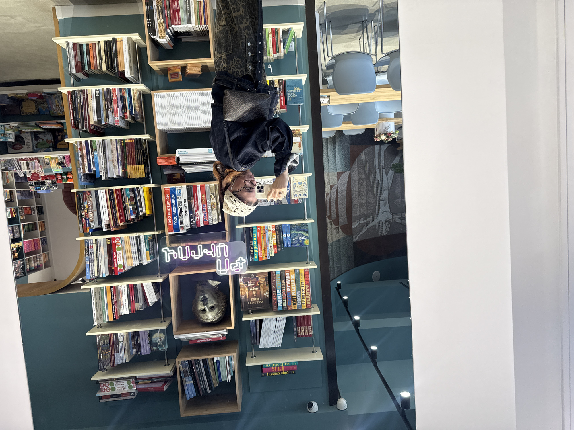 Here is a description of the image:
The image depicts a woman standing in front of a bookshelf. She is wearing a dark jacket, animal print pants, and a white head covering. She is holding a phone and appears to be taking a photo of herself or something on the bookshelf.
The bookshelf is filled with various books and objects, including a neon sign and a sculpture. The setting appears to be a bookstore or library, with other shelves visible in the background.
The image was taken in Yerevan, Armenia, based on the text and names shown on the books.