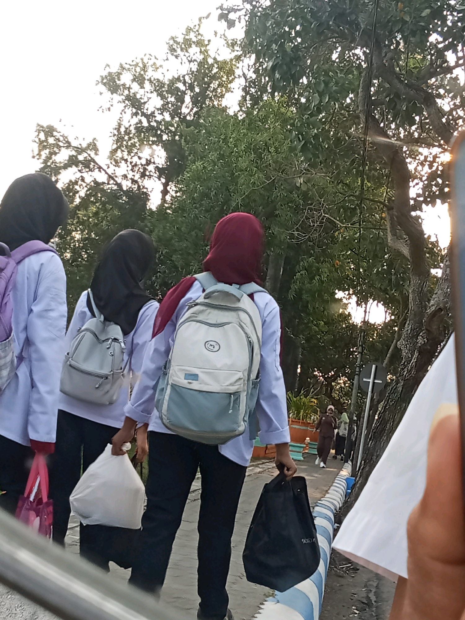 The image depicts three young women walking away from the camera on a paved path. They are all wearing white tops and dark pants, and they each have a backpack. The woman on the left has a purple backpack, the middle woman has a light gray backpack, and the woman on the right has a light gray and blue backpack. The woman in the middle is carrying a white plastic bag. The woman on the right is carrying a black tote bag. In the background, there are trees and a street with a white cross sign. There are other people in the distance walking on the street. The scene appears to be in a public area, possibly a school or a park, in the city of Kaliwungu, Indonesia. The lighting suggests it is daytime, with the sun partially obscured by trees.