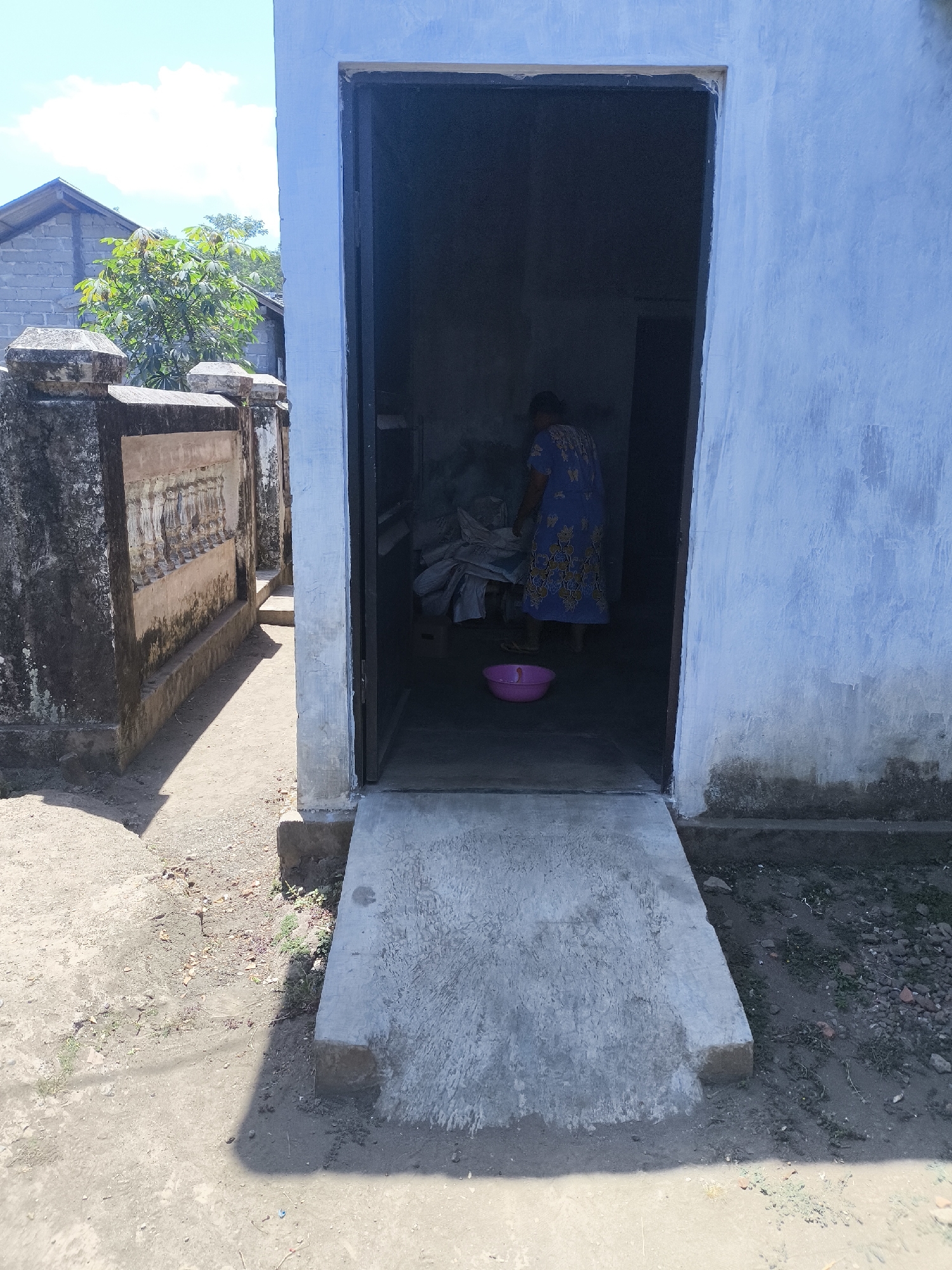 The image depicts an outdoor scene in the city of Grati, Indonesia. In the foreground, a light blue painted building wall is visible on the right, showing signs of weathering and water stains at the base. A concrete ramp extends from the building's entrance, which is an open doorway leading into a dark interior. To the left of the building, a low, weathered stone wall with decorative elements runs along a pathway. Beyond the wall, a house with a gray brick structure and a pitched roof is visible, along with green foliage. Inside the building's doorway, a person wearing a blue and yellow patterned dress is visible from the back, appearing to be engaged in some activity within the dimly lit interior. A pink plastic bowl is on the floor near the person. The ground is a mixture of dirt and gravel. The sky is clear with some clouds.