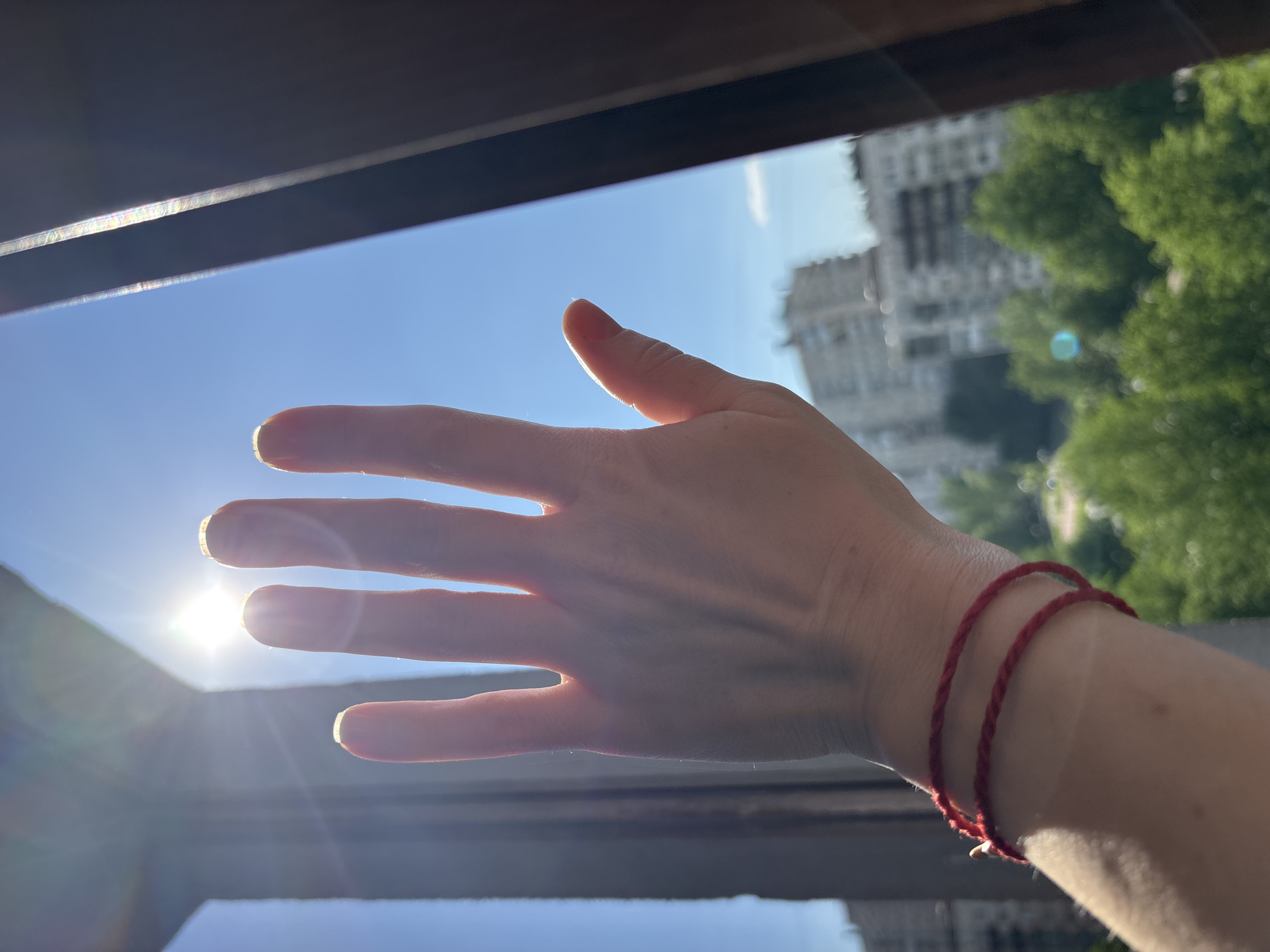 A fair-skinned human hand is positioned near a window with the sun shining through. The hand has a red string bracelet on the wrist. Outside the window is a blurred view of buildings and green trees, likely in a city. The location is Unknown, Russia. The scene appears to be shot during a sunny day.