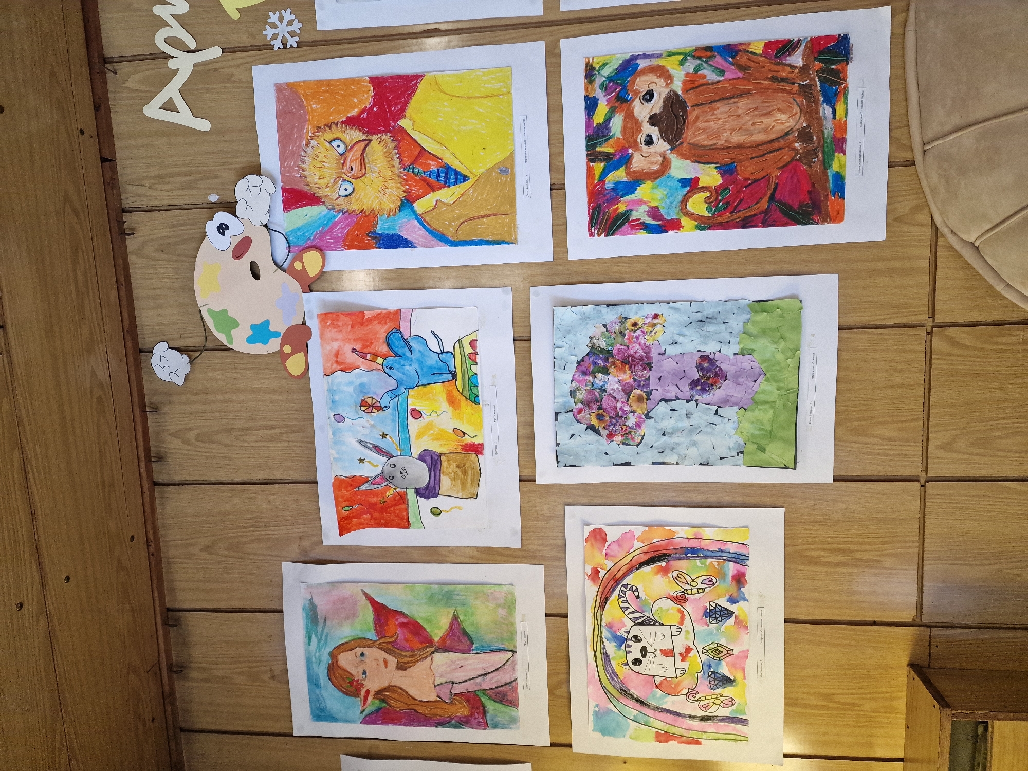 The image shows a wall with several children's drawings displayed on it. The wall is made of vertical wooden panels. Above the drawings, there is a cutout of a cartoonish art palette with eyes and arms, and to the right, the word "April" is written in large wooden letters, with a snowflake cutout nearby.
The drawings, arranged in a grid, are:
Top row, left: A drawing of a fairy with long red hair and wings.
Top row, center: A drawing of a blue elephant with a party hat, standing in a box, with balloons in the background.
Top row, right: A drawing of a bird with yellow feathers and an angry expression, wearing a yellow jacket and a blue tie.
Bottom row, left: A drawing of a cat with stripes on its face, sitting on a cloud, with a rainbow behind it and butterflies.
Bottom row, center: A mosaic-style artwork of a vase filled with colorful flowers, with a green base.
Bottom row, right: A drawing of a monkey sitting on a branch, with a colorful, abstract background.
Below each drawing, there are small white labels with text, likely indicating the title of the artwork and the artist's name.
In the bottom left of the image, a corner of a wooden cabinet or shelf is visible. The overall setting appears to be a classroom or a hallway where children's artwork is displayed.